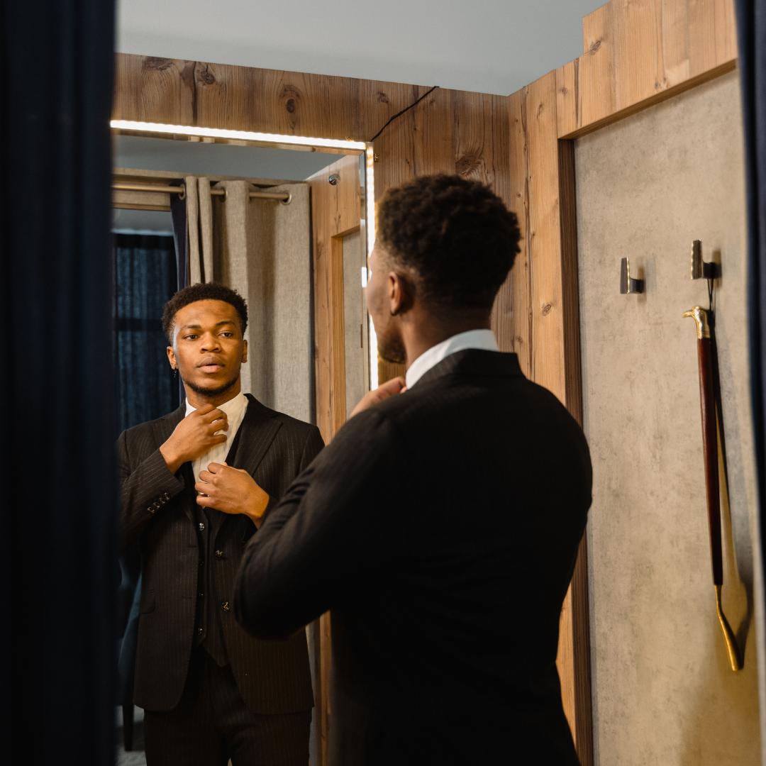 A man in a suit adjusts his tie while looking at himself in a mirror, preparing for a formal event or photoshoot.