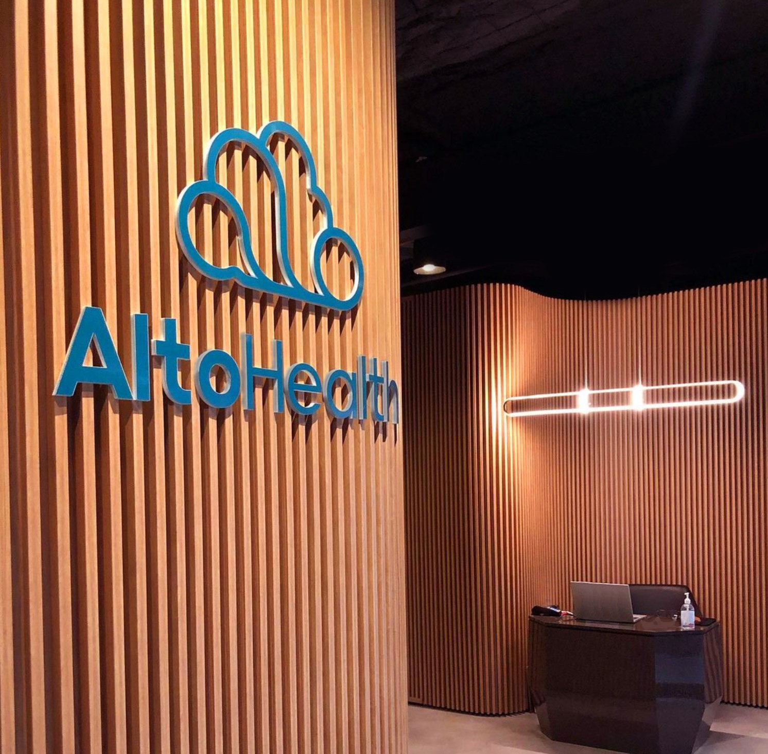 Freelance wayfinding design and signage for Alto Health, the logo is highlighted by lighting on warm timber paneling in the entrance to the business