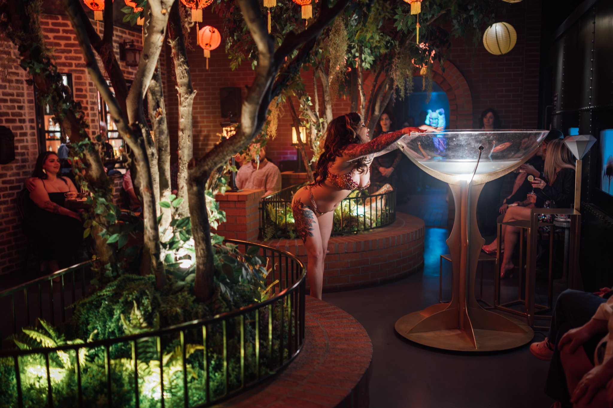 A woman with long red hair and tattoos on her arms, dressed in a glittery top, leans over a transparent water-filled sculpture in an indoor space with brick walls and hanging lanterns, while several people seated around observe her.