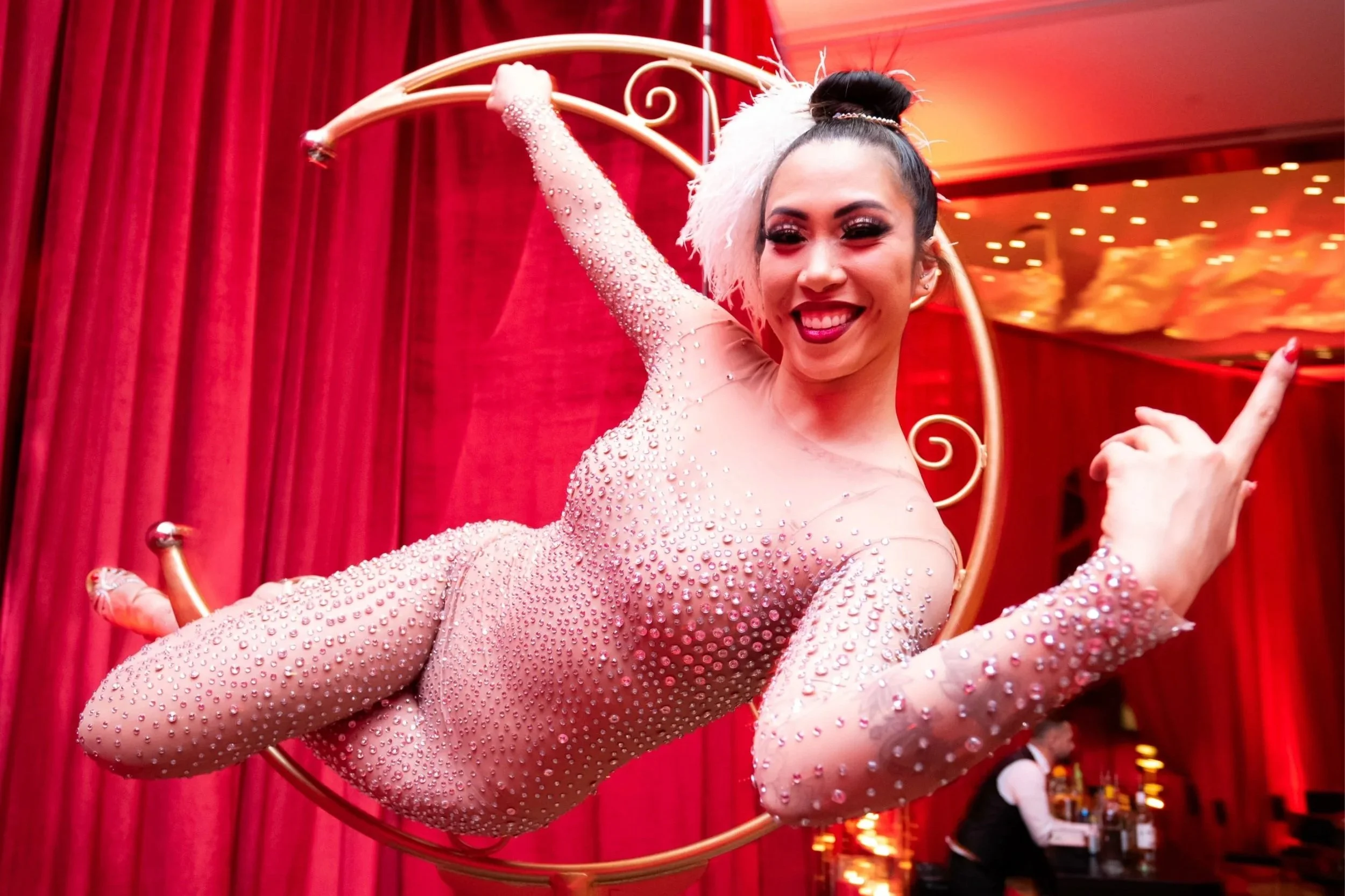 A smiling woman in a pink sparkling outfit performing an aerial hoop act in front of red curtains.