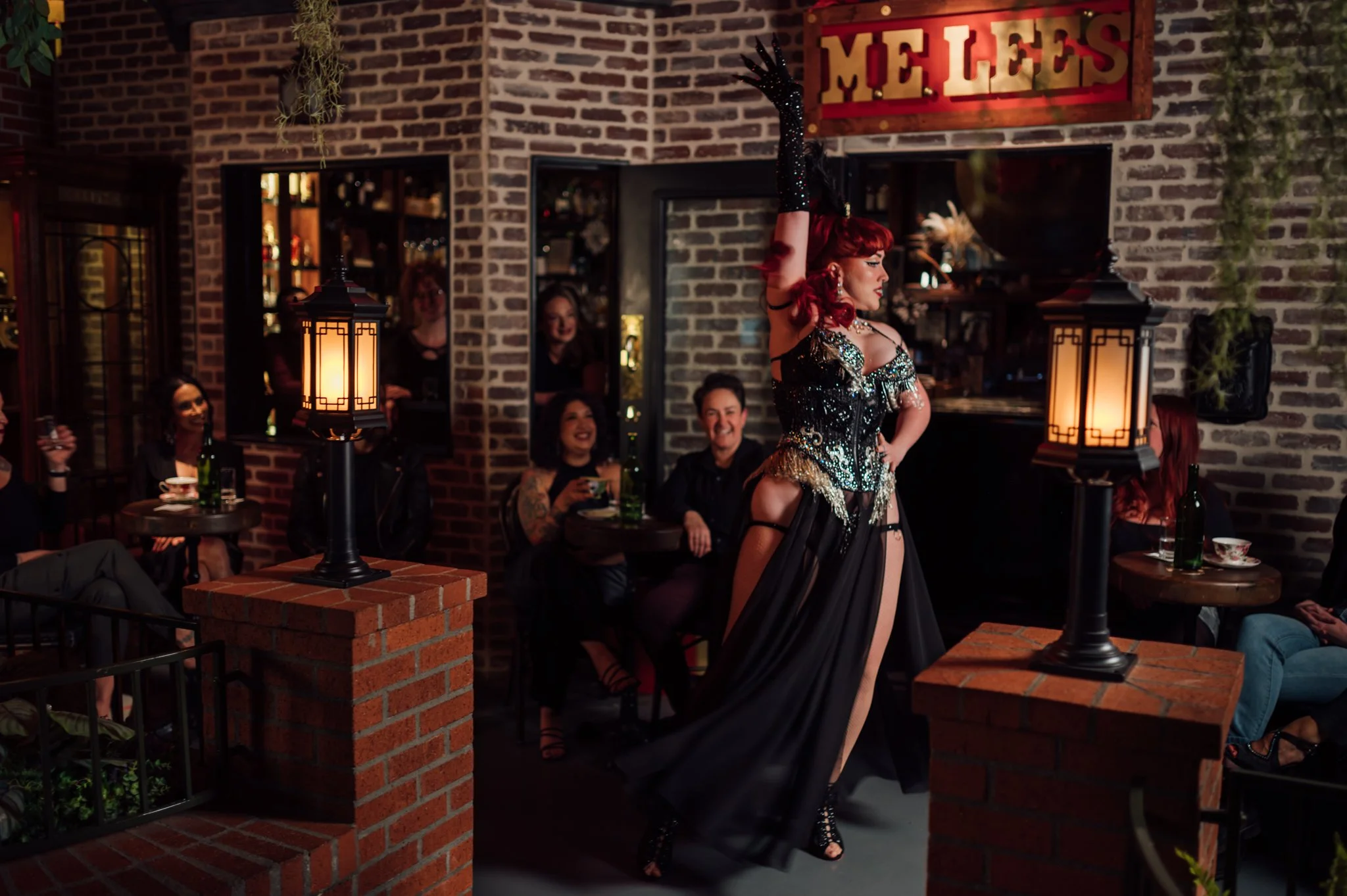 A performer with red hair, wearing a black and gold costume with high slit, performs on stage in a restaurant or bar with brick walls. Audience members are seated at tables, watching and smiling. The setting features warm lighting and a sign that reads 'Mel's'.