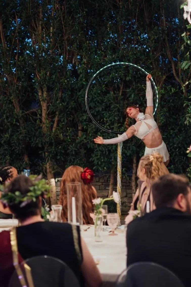 A performer is hoop dancing during a nighttime outdoor event, with an audience seated at tables watching.