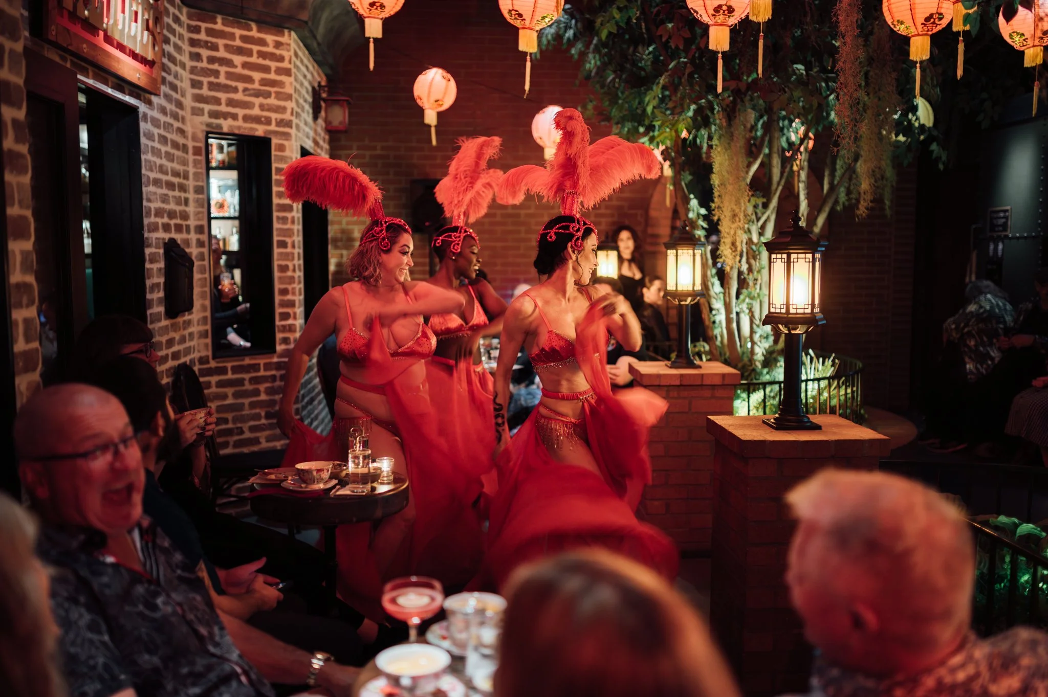 Performers in red costumes with large pink feathered headpieces dancing inside a restaurant with exposed brick walls, lanterns, and hanging plants, while seated guests watch.