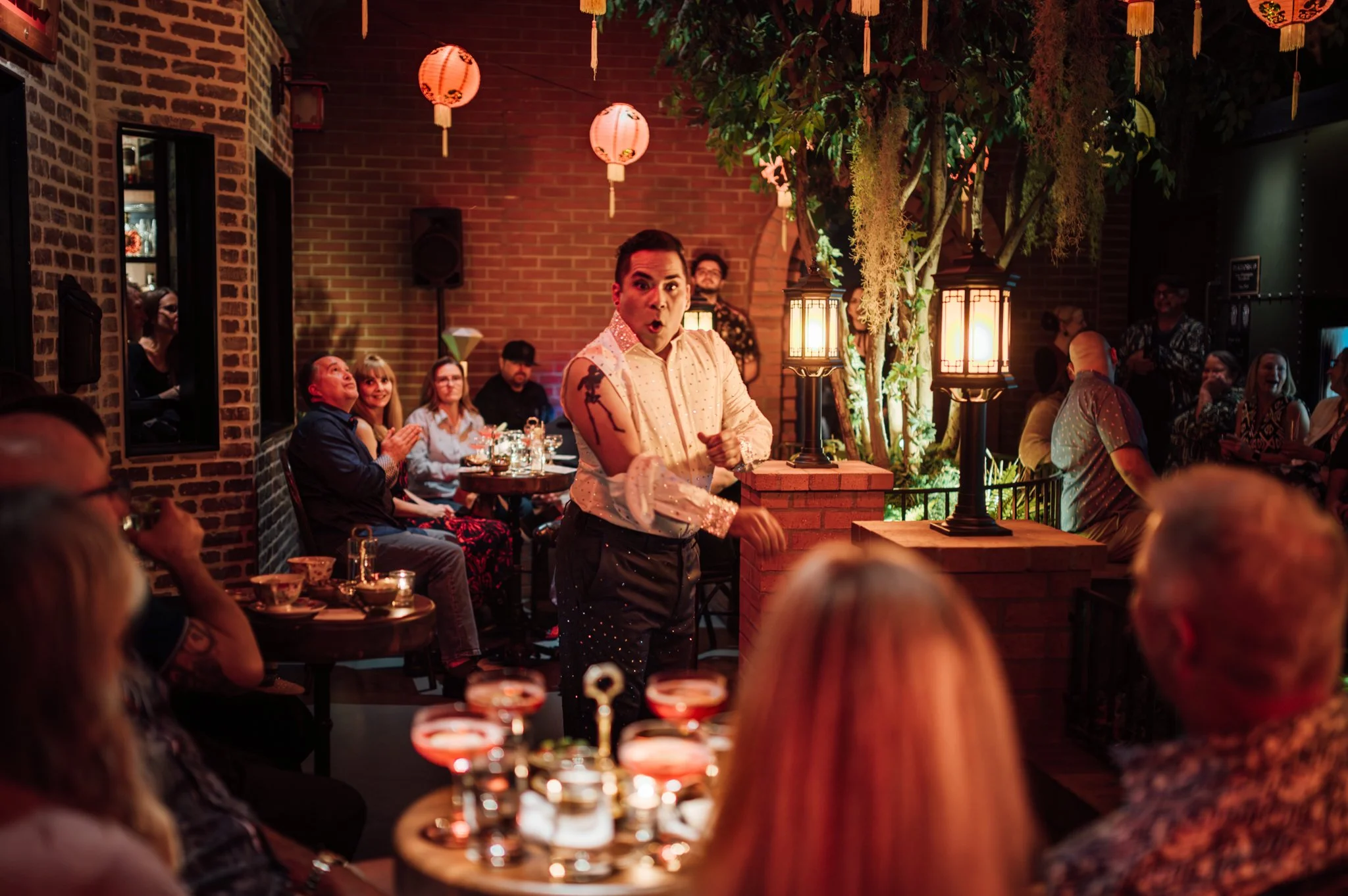 A lively indoor restaurant or event space decorated with hanging Japanese paper lanterns and large ornamental plants. A performer with a surprised expression stands in front of a seated audience clapping and enjoying the entertainment.