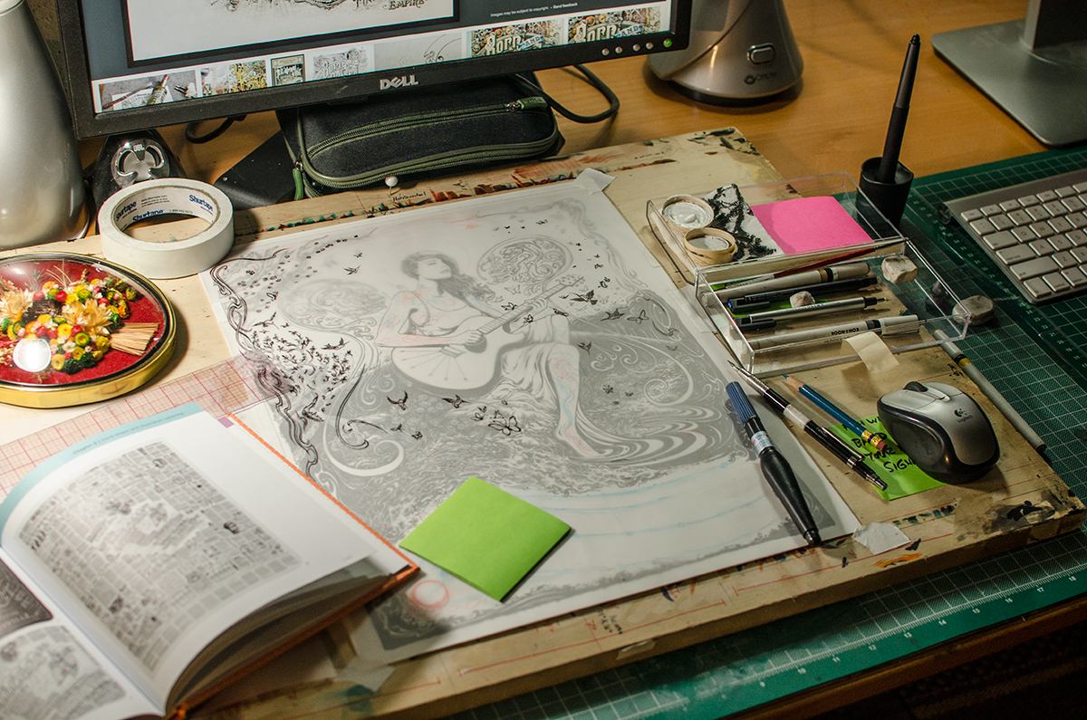 Studio work desk, cluttered with art supplies, computer equipment, various reference books, and a flower talisman . The secondary vellum layer I used to draw extra details and place the text is also visible.