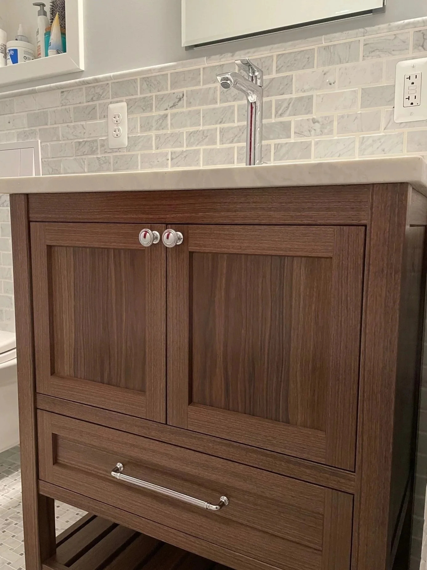 A wooden bathroom vanity with a white stone countertop, a chrome faucet, and a shelf with toiletries above it. The wall behind is covered with small white marble tiles, and there are two electrical outlets visible.