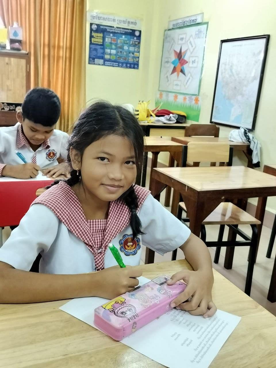 Smiling young girl in school uniform sitting at a wooden desk, holding a green pen and working on a worksheet with a pink pencil case featuring cartoon characters, in a classroom with another student and educational posters on the walls.