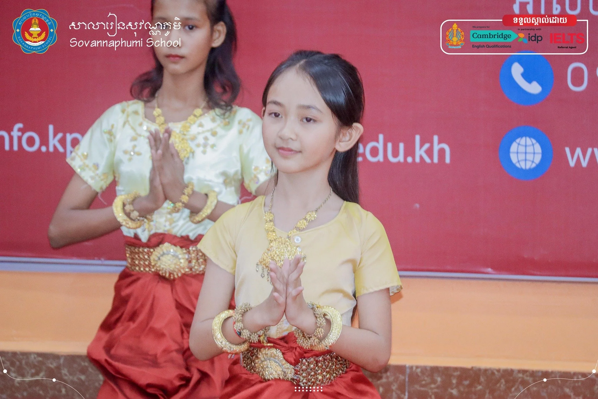 Two young girls dressed in traditional Cambodian attire, sitting with hands pressed together in a greeting gesture, in front of a red backdrop with school and educational logos.