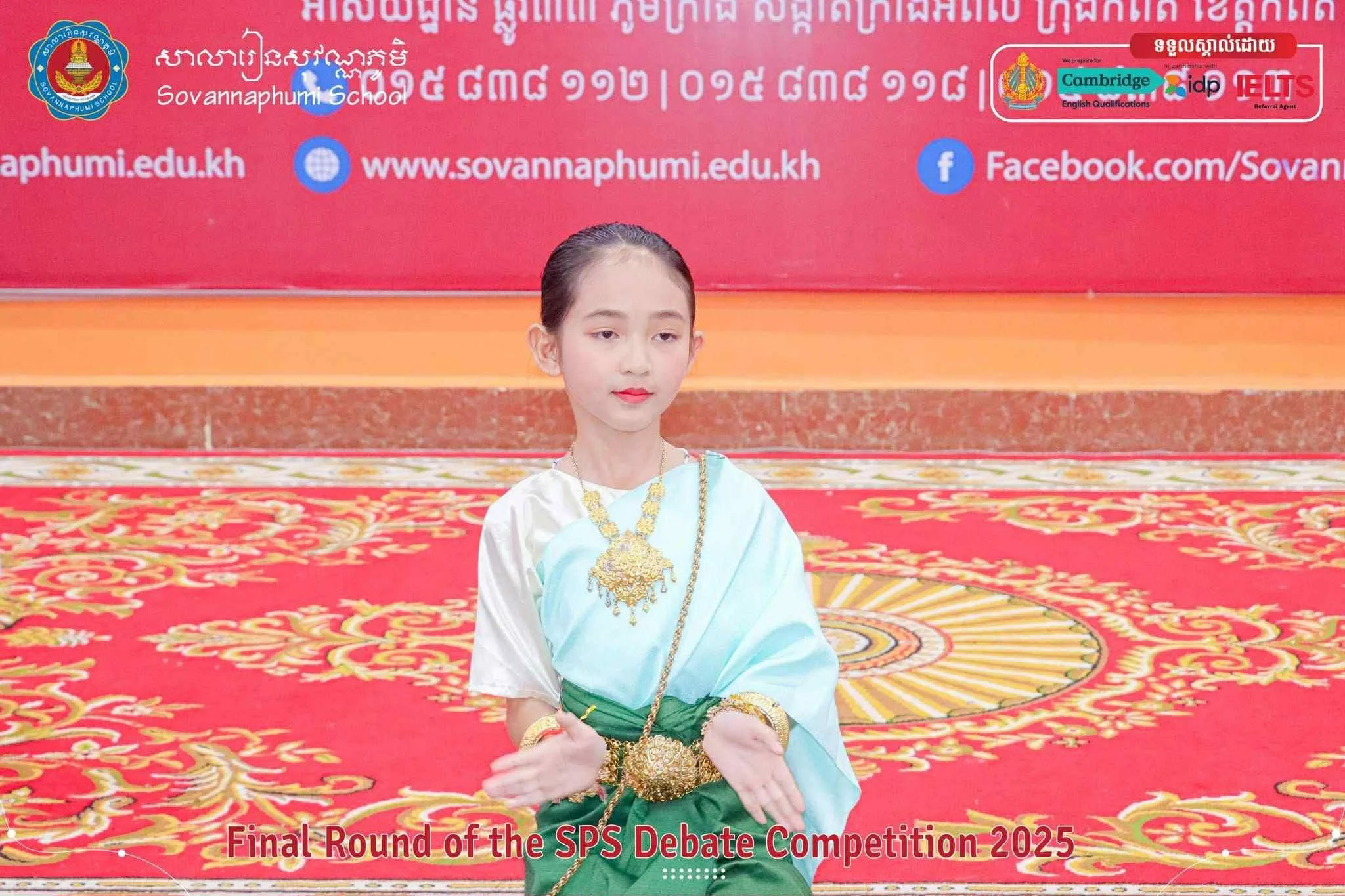 A young girl dressed in traditional attire sitting with her hands crossed on a vibrant red and gold carpet during the final round of the SPS debate competition 2025 at Sovannaphumi School.
