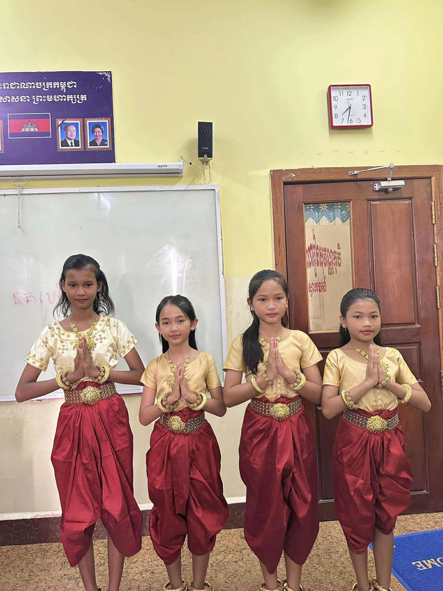 Four young girls in traditional Cambodian dance costumes, standing indoors with their hands in prayer position.
