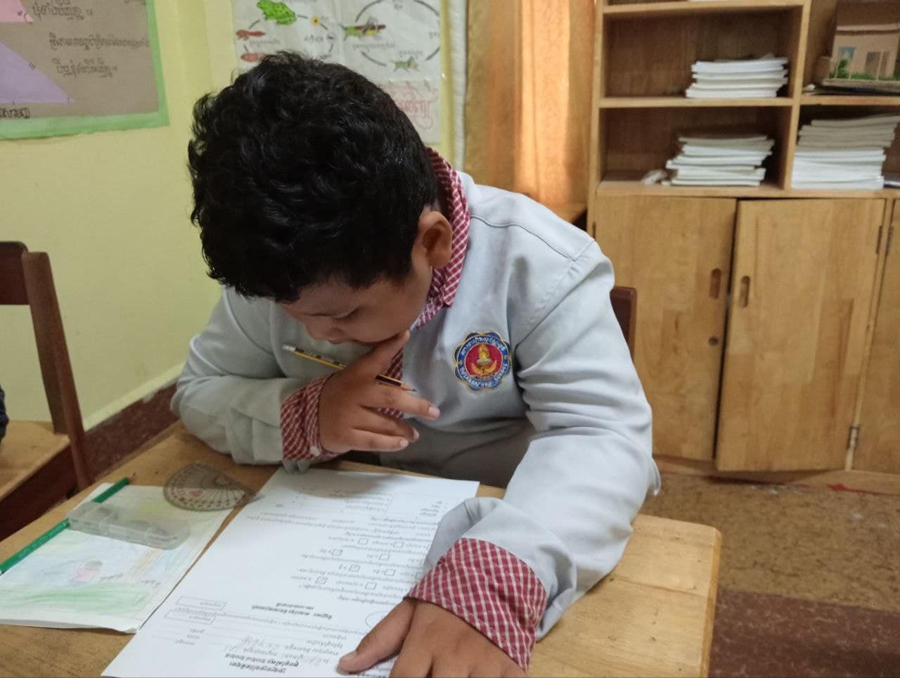A young student sitting at a wooden desk in a classroom, leaning over a worksheet with a pencil in hand, wearing a white uniform with red checkered cuffs and a school emblem.