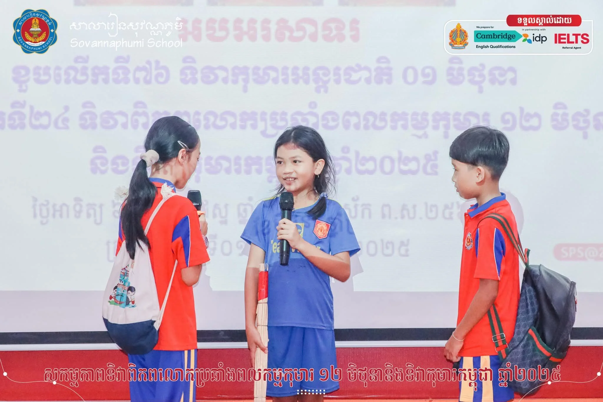 Three children standing in front of a presentation screen, engaged in a conversation. Two girls and one boy, all wearing school uniform backpacks and holding microphones. The girl in the middle is smiling while speaking. The background features text and logos, indicating a school event or presentation