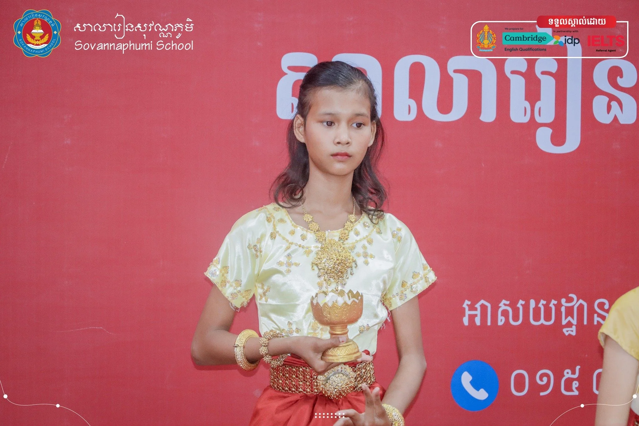 A young girl dressed in traditional attire holding a small golden bowl, standing in front of a red backdrop with text and logos, including Sovannaphumi School and IELTS.