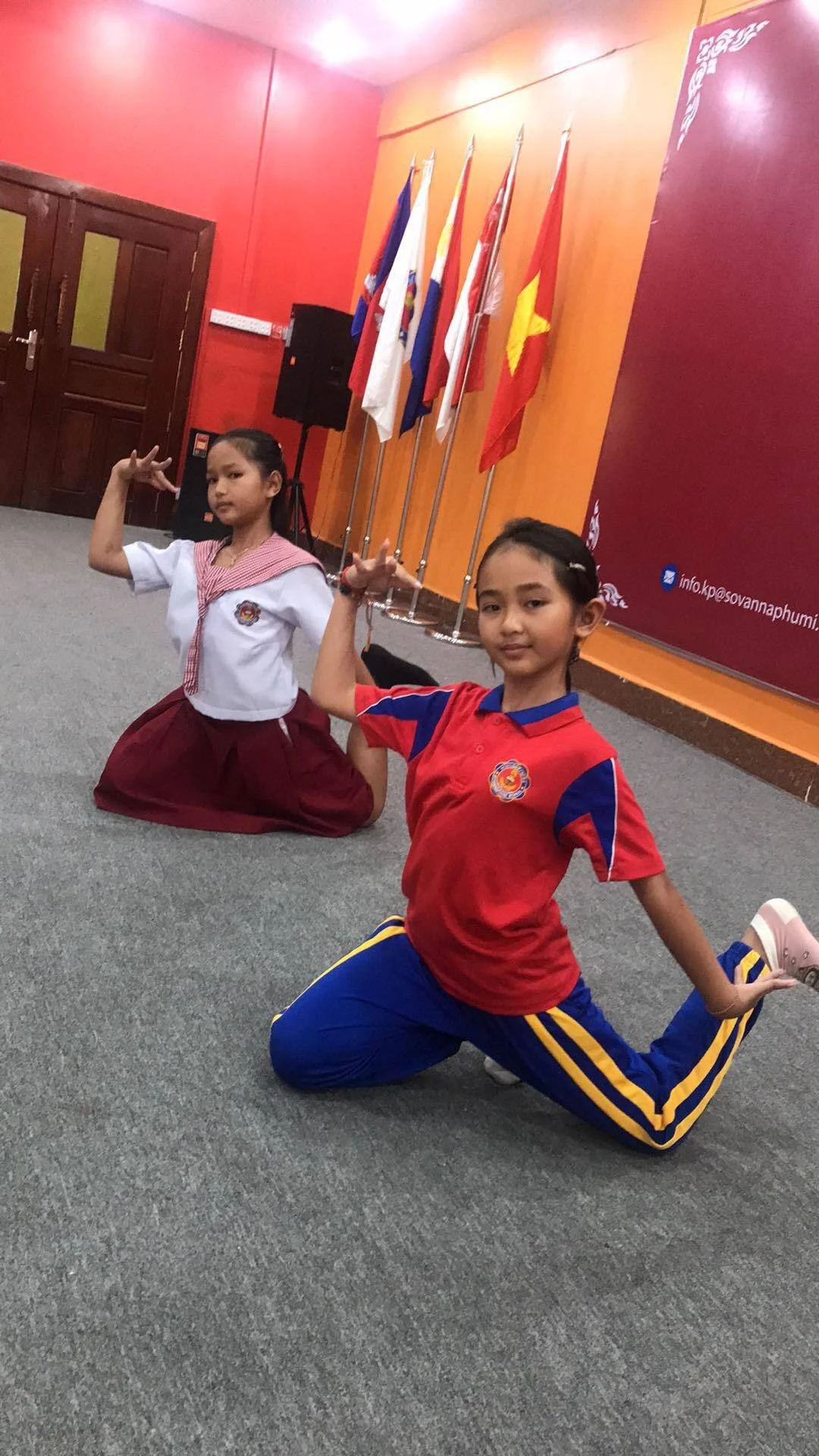 Two young girls in school uniforms performing yoga poses on a grey carpeted floor in front of flags and a red wall.