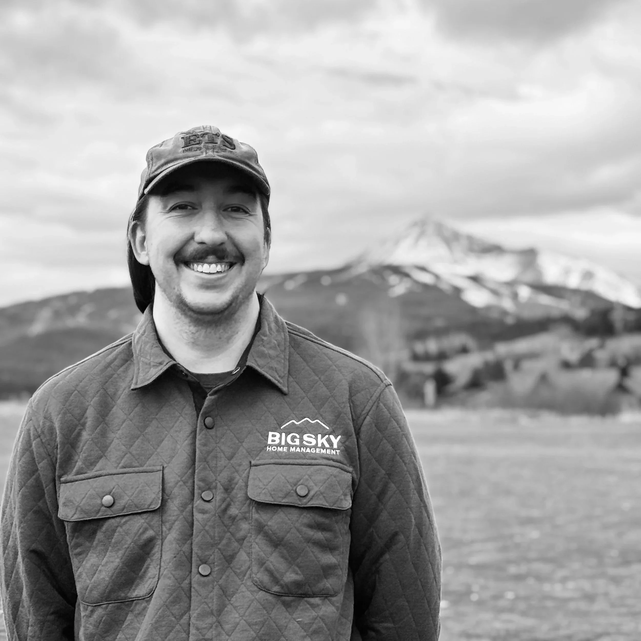 A smiling man wearing a baseball cap and a quilted work jacket with a logo that says 'Big Sky Home Management' stands outdoors in front of a mountain and cloudy sky.