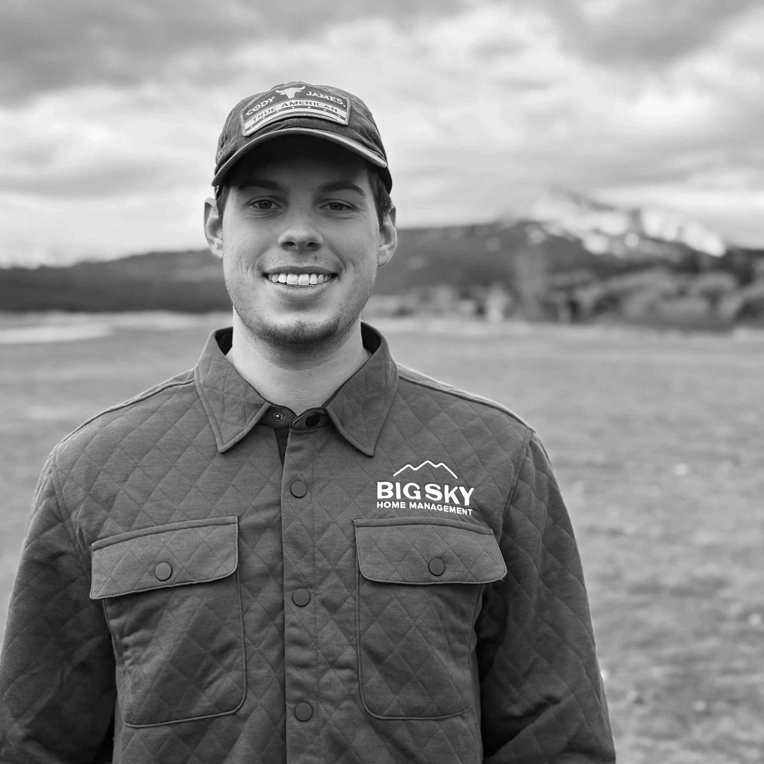 A smiling young man wearing a cap and a quilted jacket with the logo 'BIG SKY HOME MANAGEMENT' stands outdoors on a cloudy day with mountains and open landscape in the background.