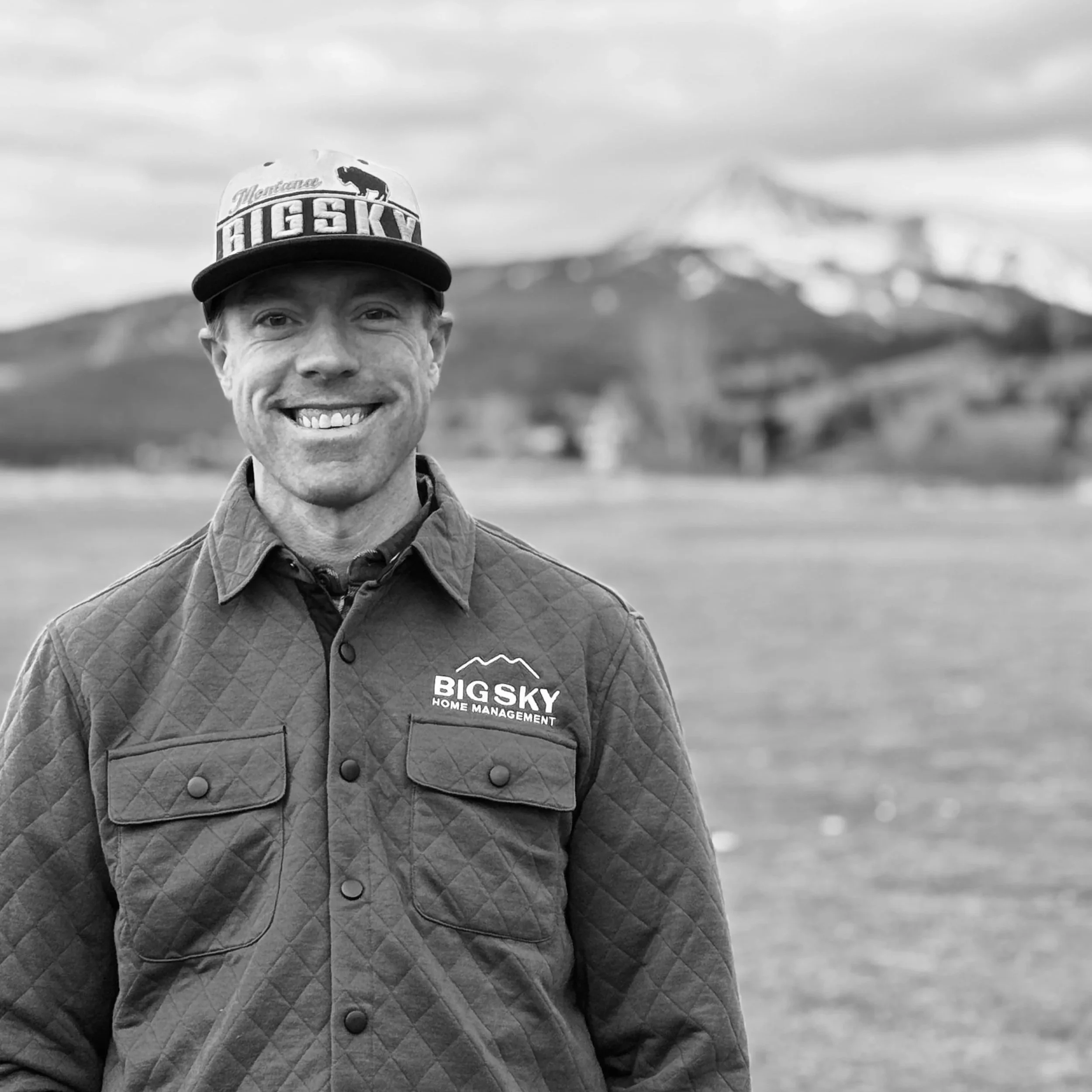 A smiling man wearing a quilted jacket and a cap with the logo 'Big Sky Home Management' stands outdoors with mountains and a snow-capped peak in the background