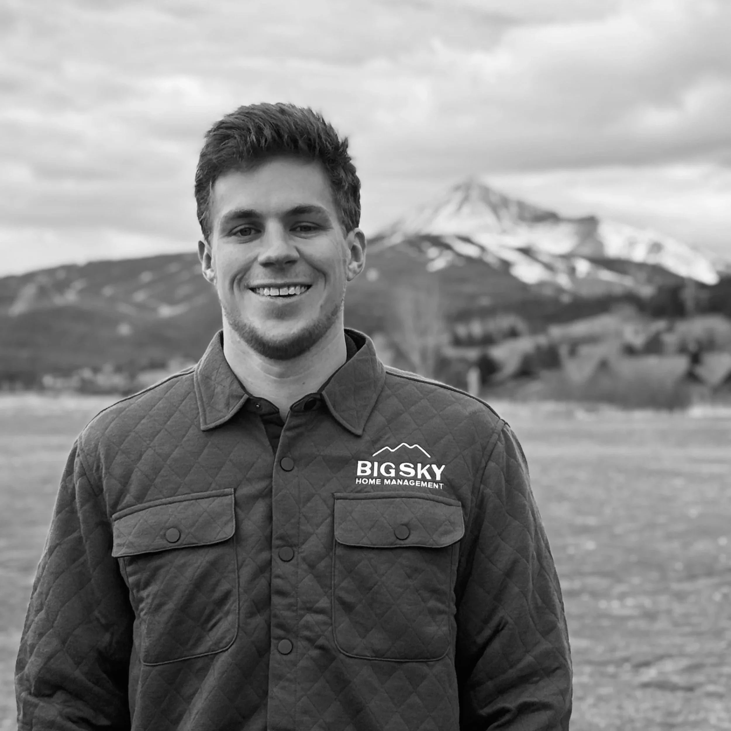 A smiling young man standing outdoors with mountains in the background, wearing a quilted jacket with a 'Big Sky Home Management' logo.