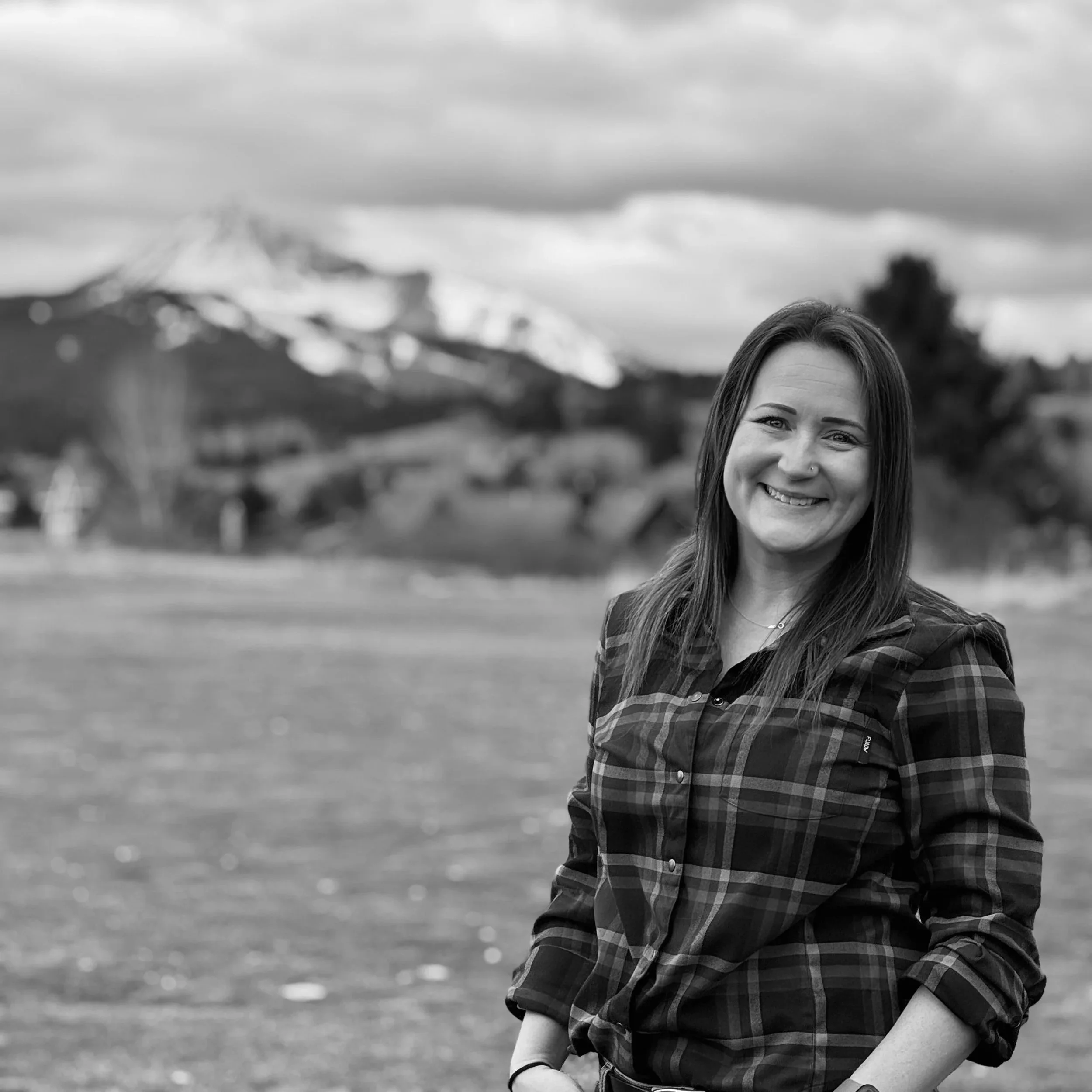 A woman with long dark hair smiling outdoors in front of a mountain landscape, snow-capped mountain in the background, overcast sky.