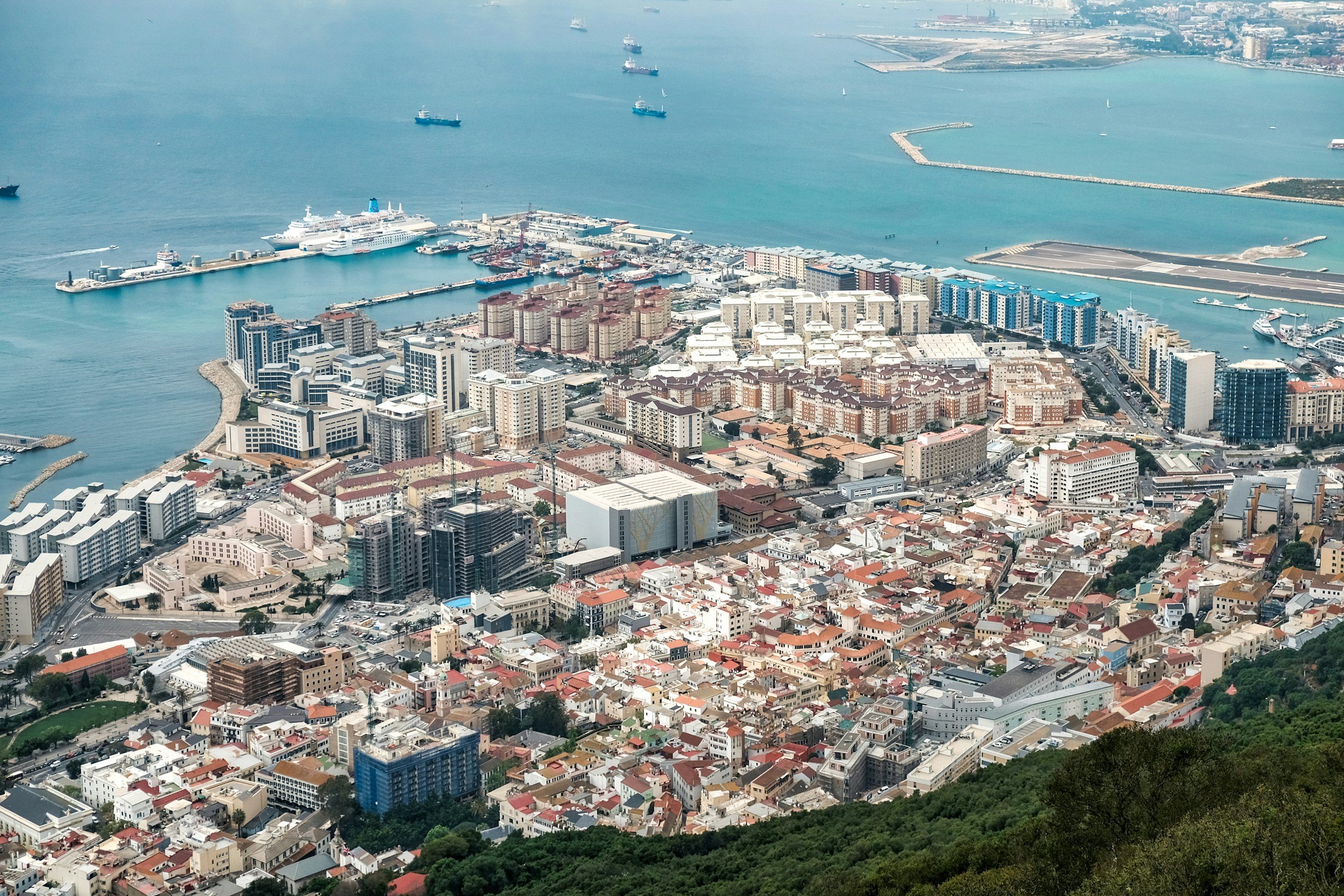 Aerial view of a coastal city with a harbor filled with ships and boats, high-rise buildings near the shoreline, and a mix of residential and commercial structures extending inland, with hills and greenery in the foreground.