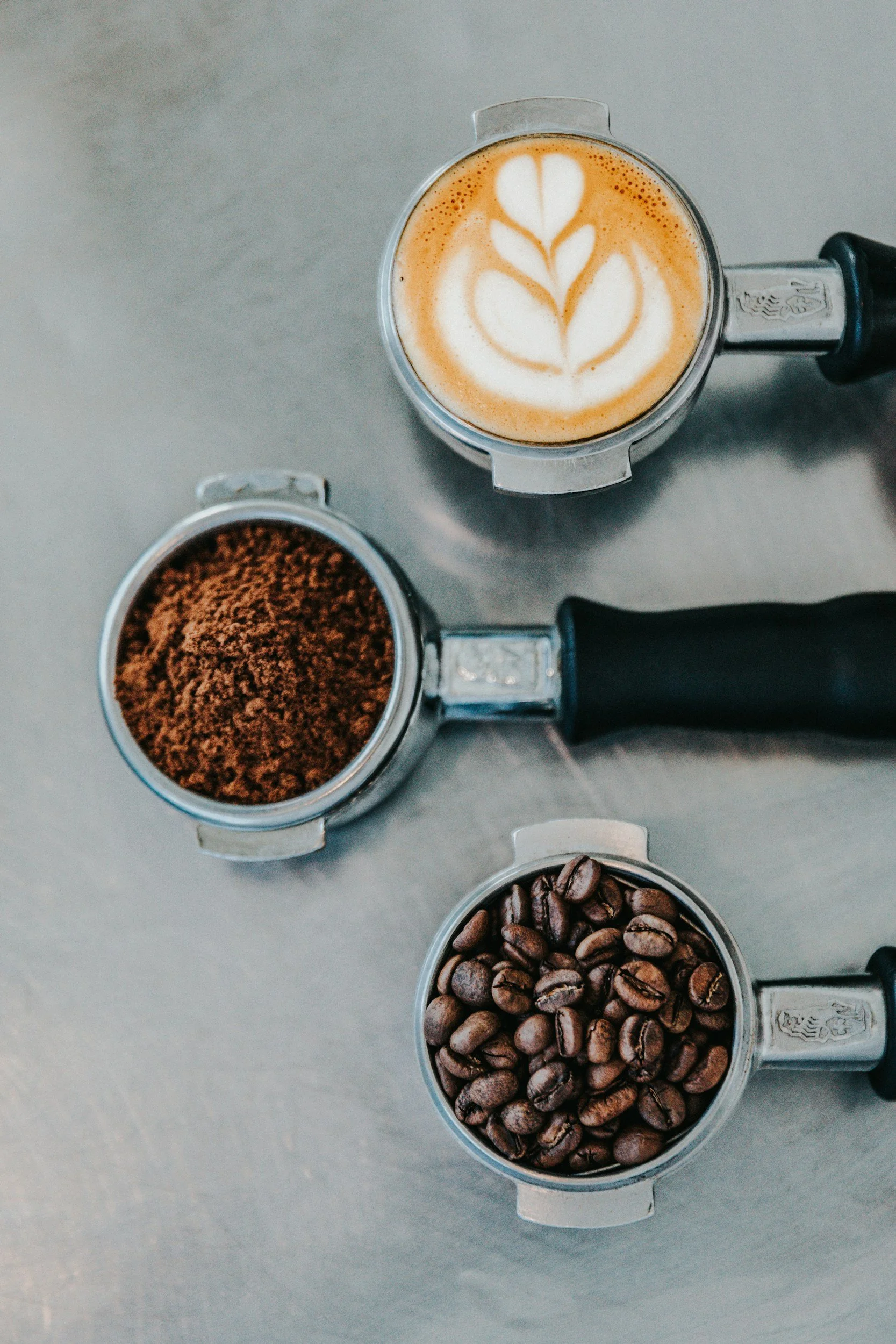 Top-down view of three stainless steel coffee portafilters filled with ground coffee, coffee beans, and a latte with latte art in a glass pitcher.