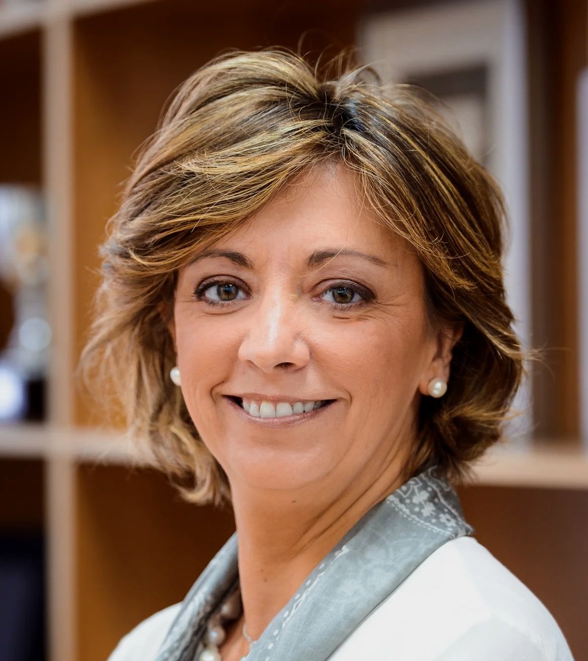 Close-up of a smiling middle-aged woman with short, wavy hair, pearl earrings, and a pearl necklace, standing in front of a wooden background.