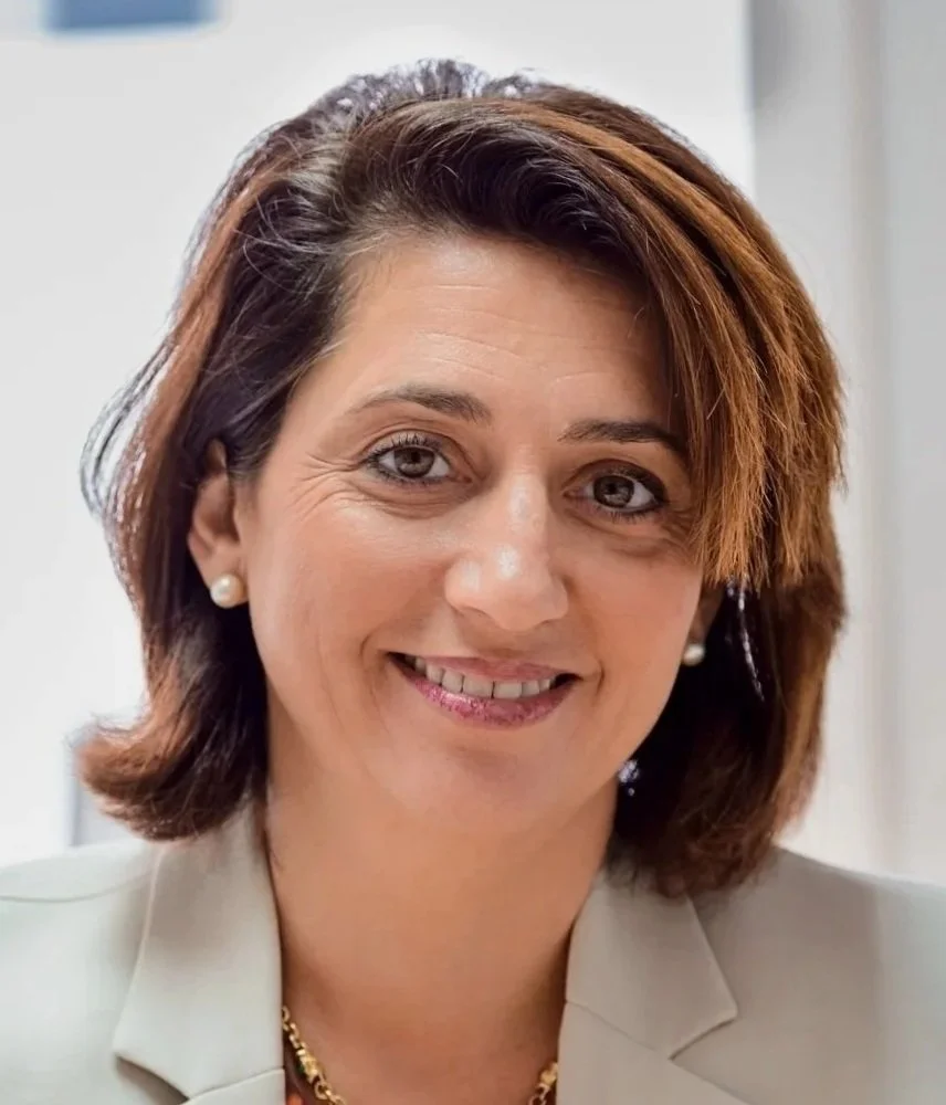 A smiling woman with short, brown hair and pearl earrings, wearing a beige blazer, in an indoor setting.