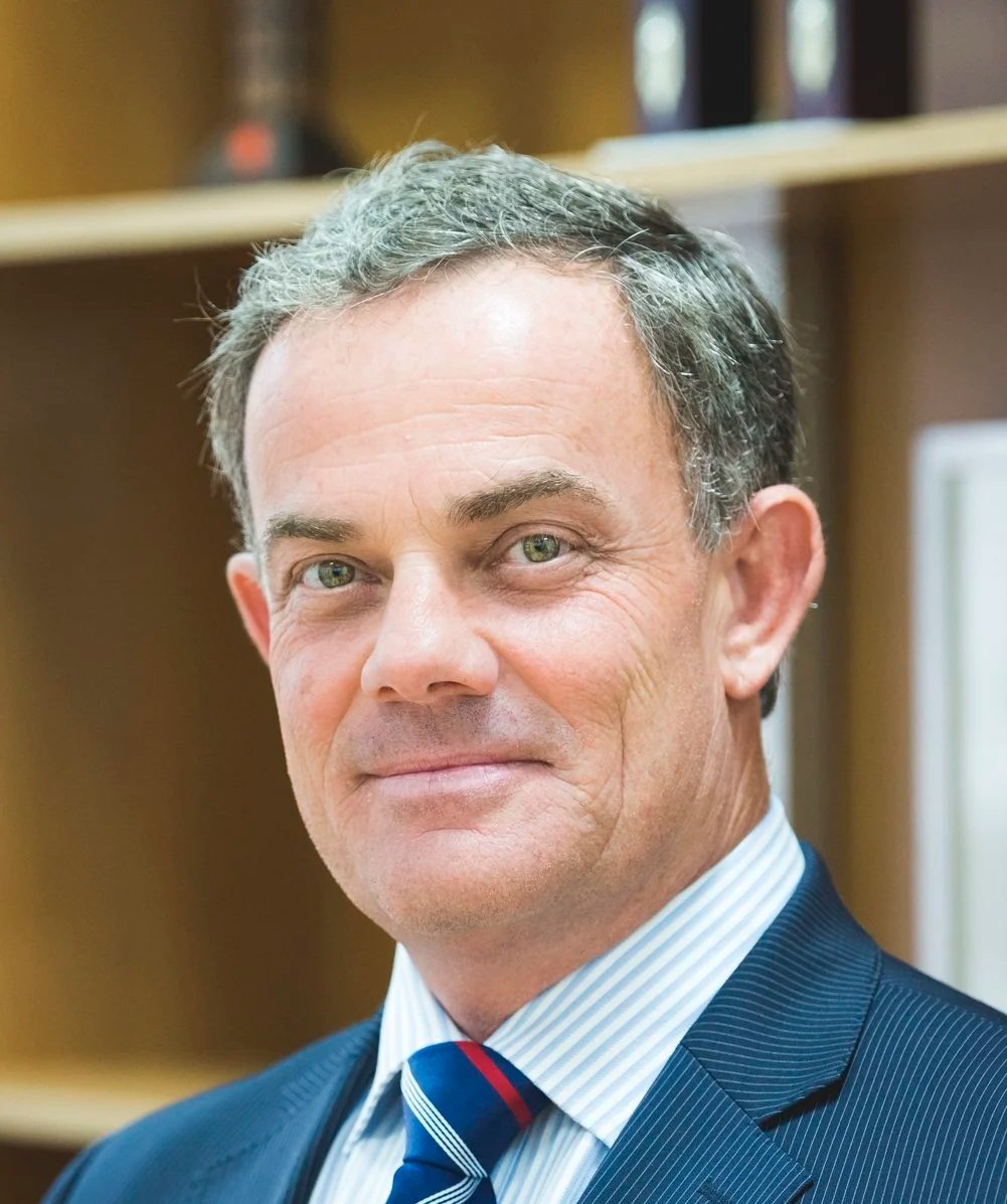 Close-up of a middle-aged man with gray hair, wearing a dark pinstripe suit, white dress shirt, and a striped blue, red, and white tie, smiling slightly in an indoor setting with wooden shelves in the background.