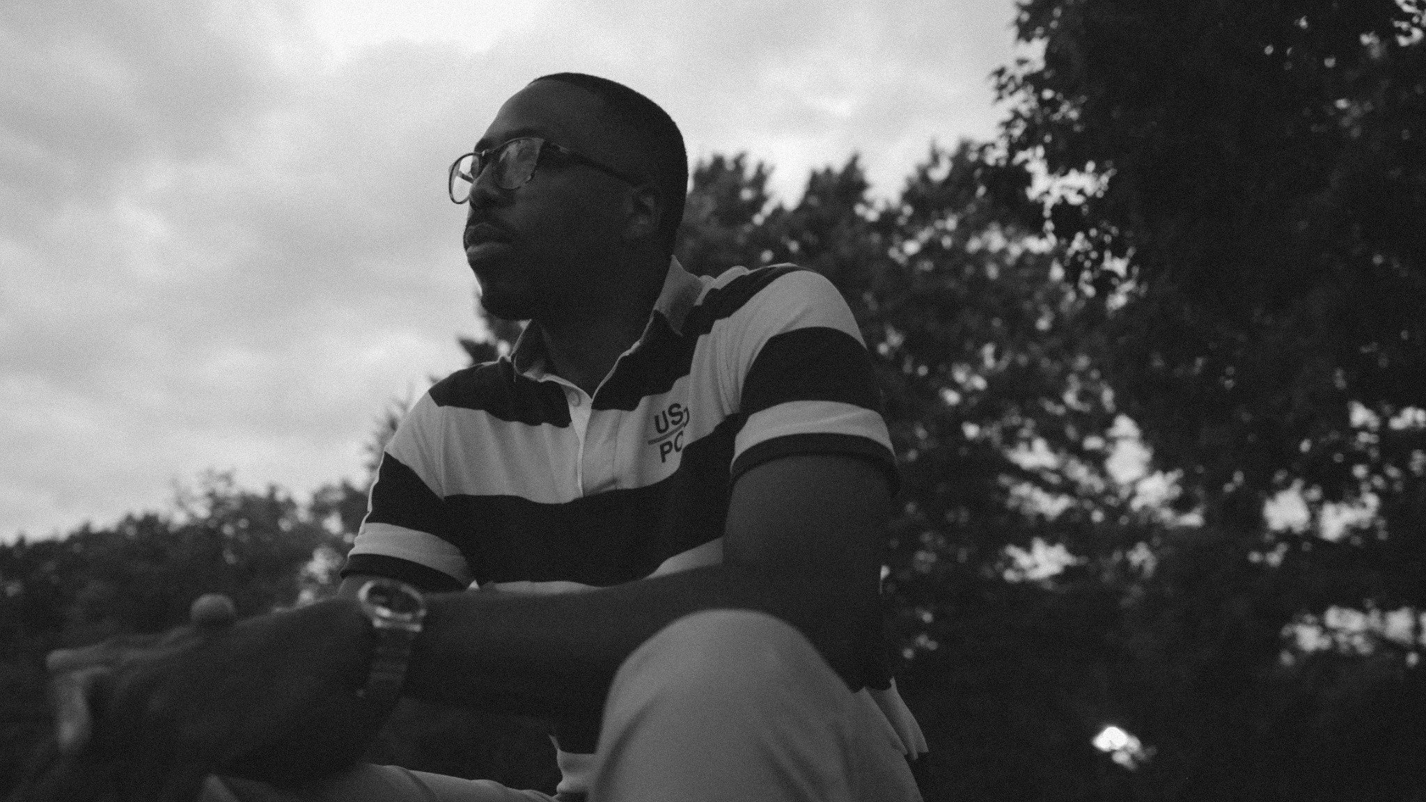 Black-and-white still of Jackson Reese seated outdoors in a striped Polo shirt