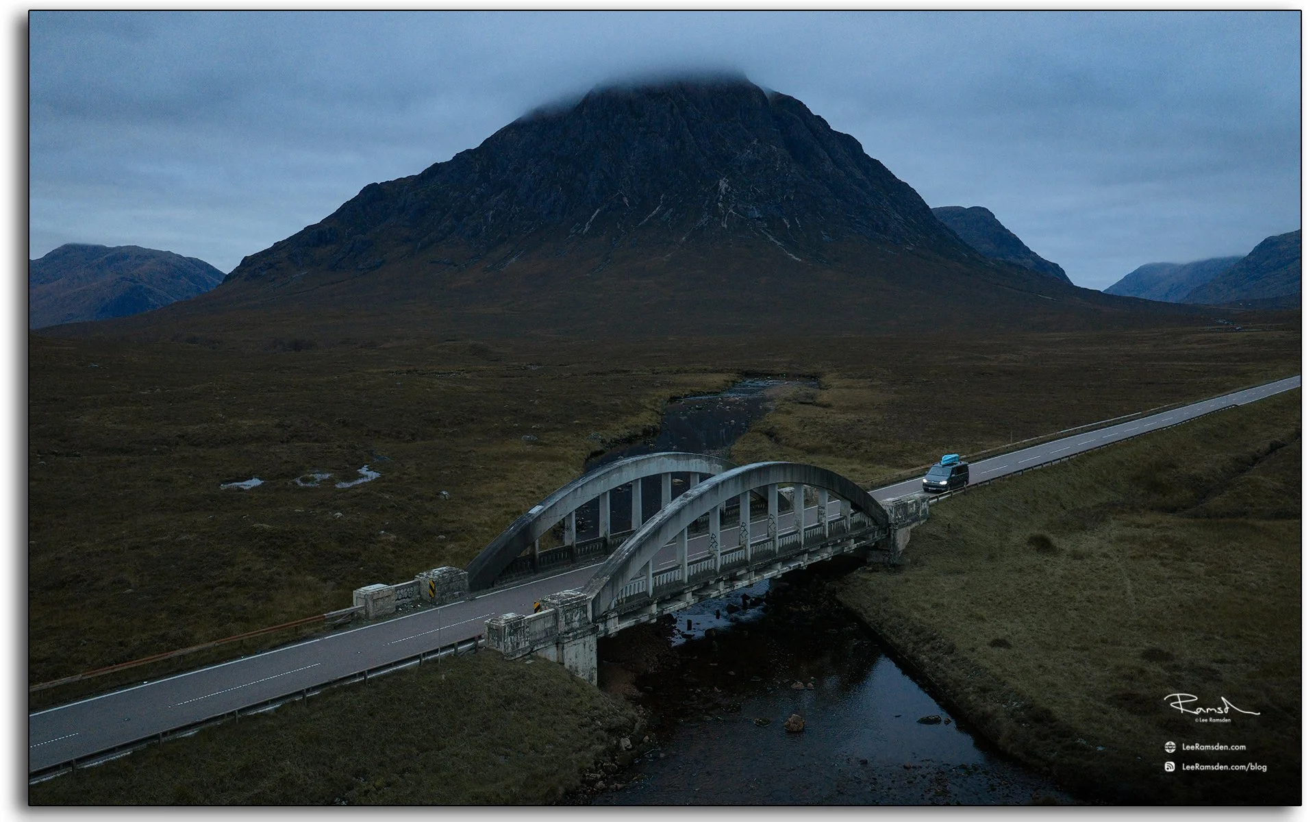 Evening aerial view of the Buachaille Etive Mòr mountain and bridge in Glencoe, Scotland, with low clouds wrapping around the peak and a van crossing the road