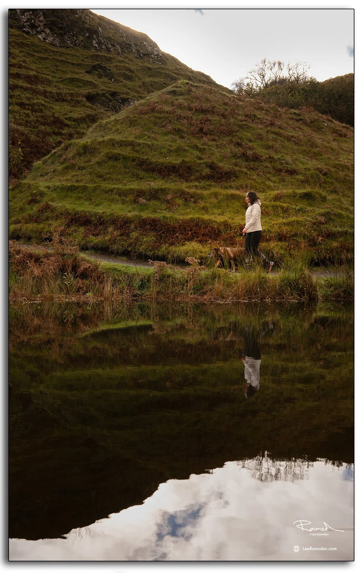Woman walking beside a calm pond at the Fairy Glen on the Isle of Skye, with her reflection mirrored in the still water and grassy hills behind