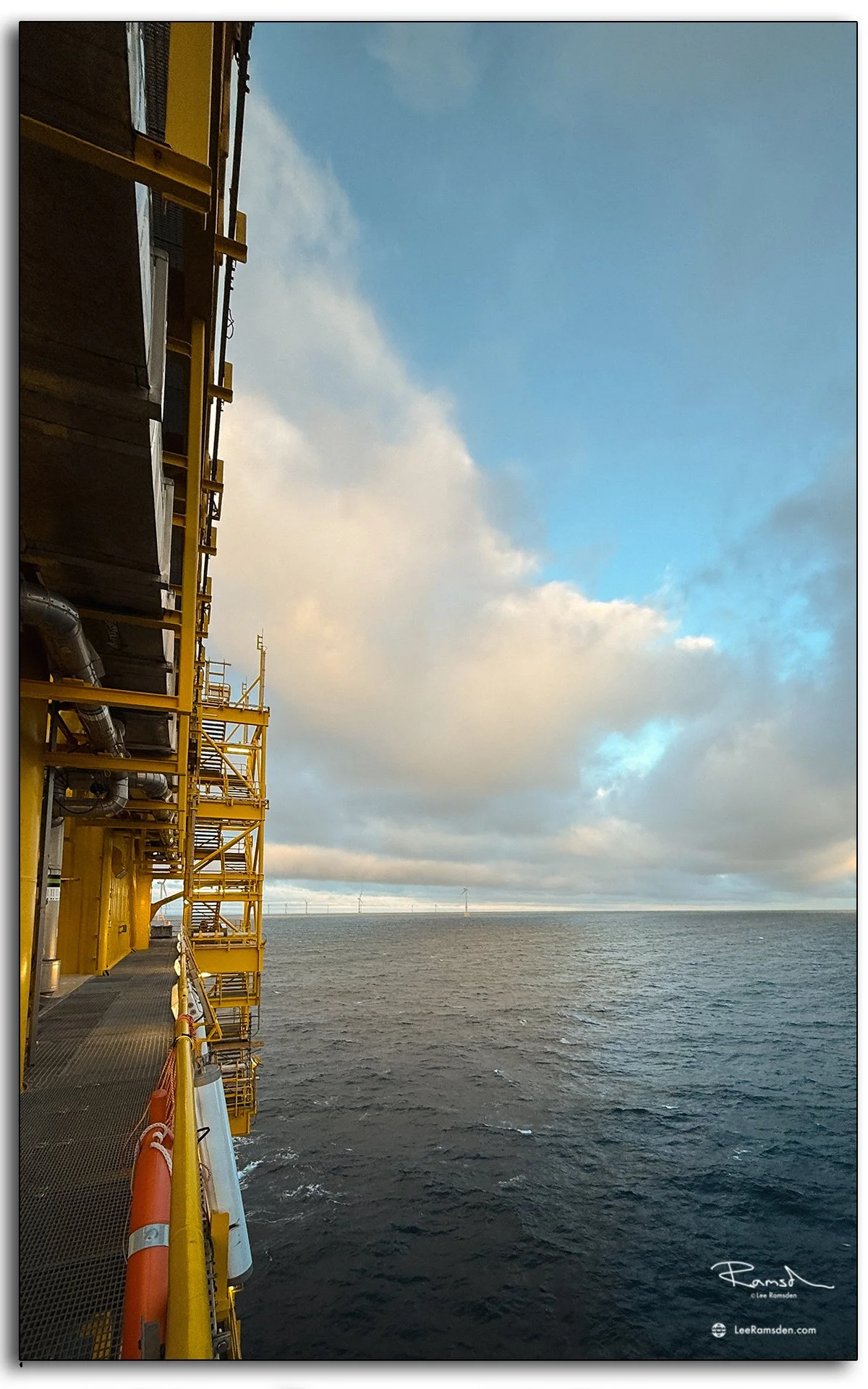 View from the side of an offshore substation platform, showing the yellow framework against a backdrop of sea and clouds.