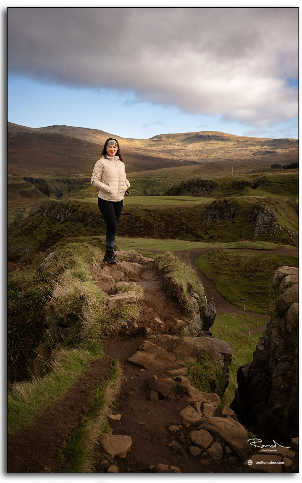 Woman standing on a rocky path overlooking the Fairy Glen on the Isle of Skye, Scotland, with sunlight highlighting the hills behind.