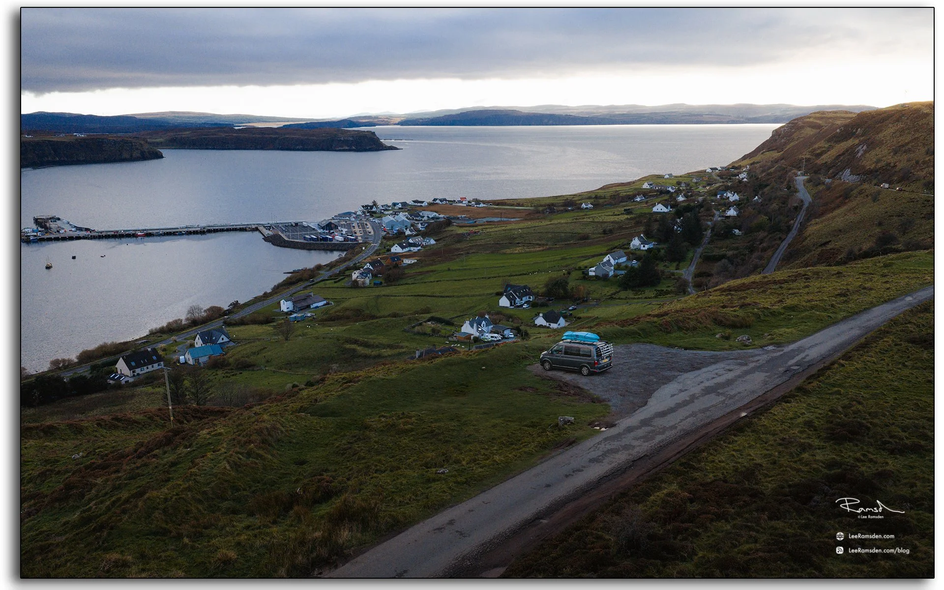 Aerial view overlooking Uig Harbour and village on the Isle of Skye, Scotland, with a camper van parked on the hillside and calm evening light over the bay