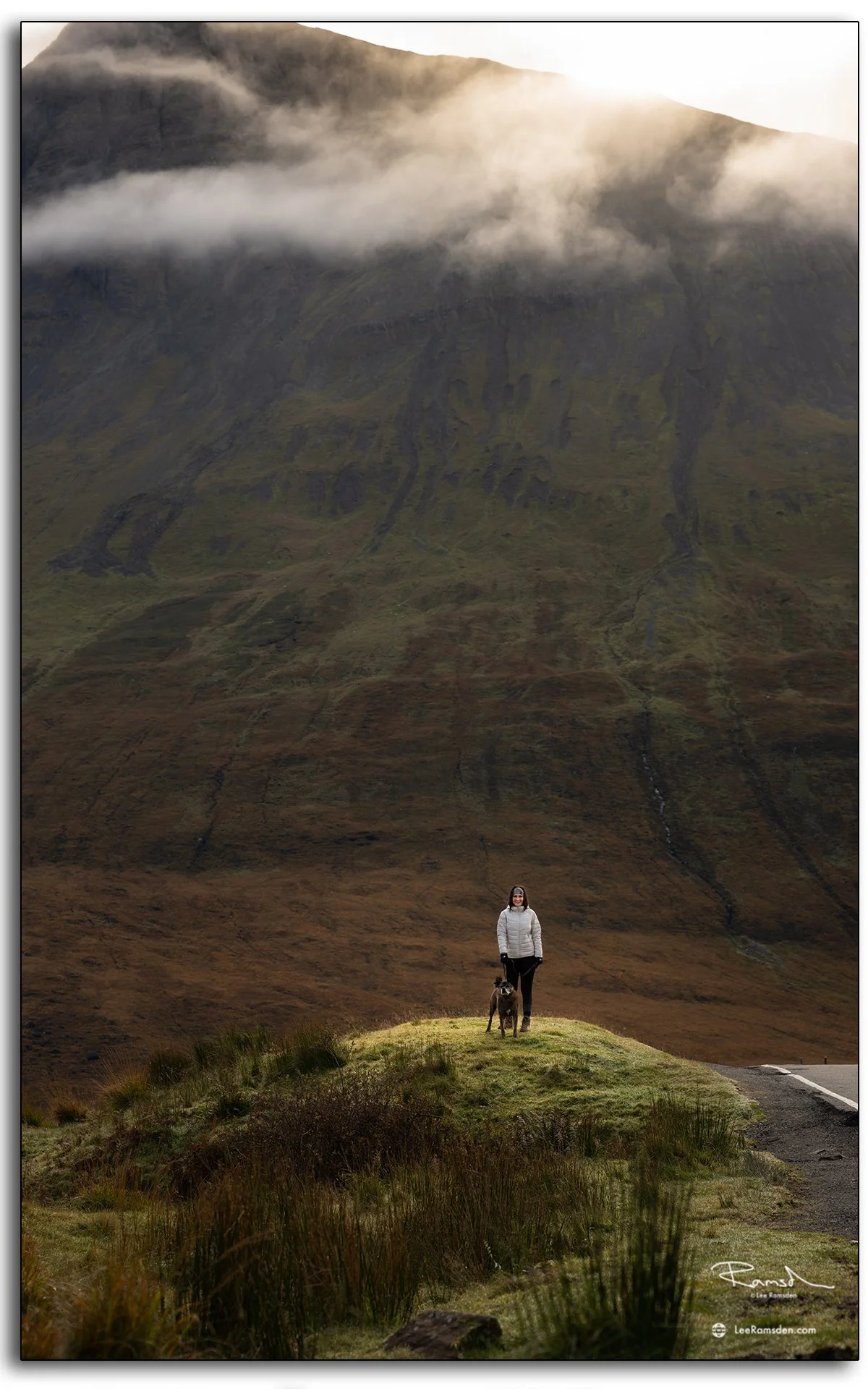 Woman standing with her dog and the foot of a rugged mountain, at the Fairy Pools on the Isle of Skye, Scotland, surrounded by golden light and rugged hills