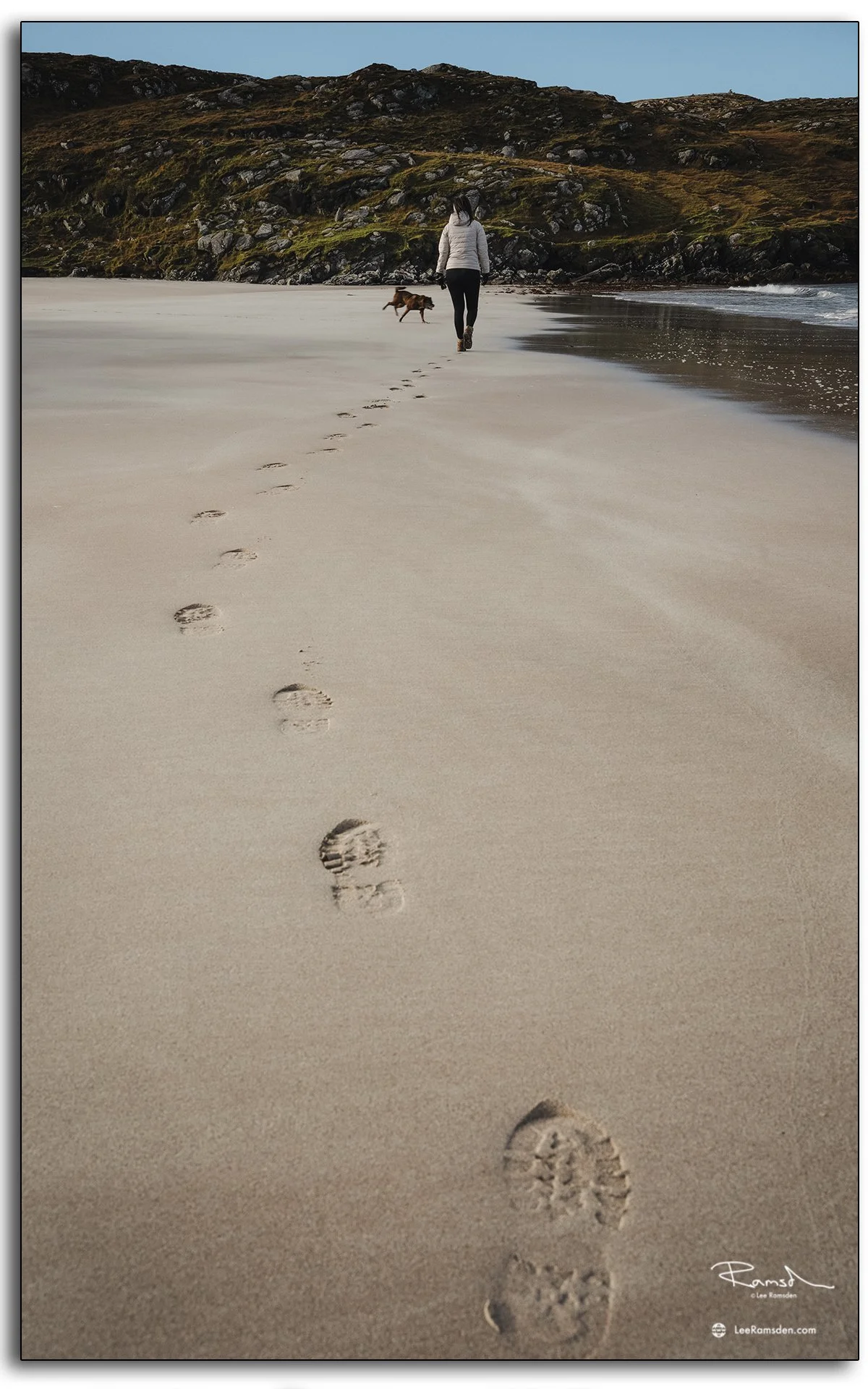 Woman walking with her dog along Hushinish Beach on the Isle of Harris, Scotland, leaving footprints in the smooth sand on a calm sunny day