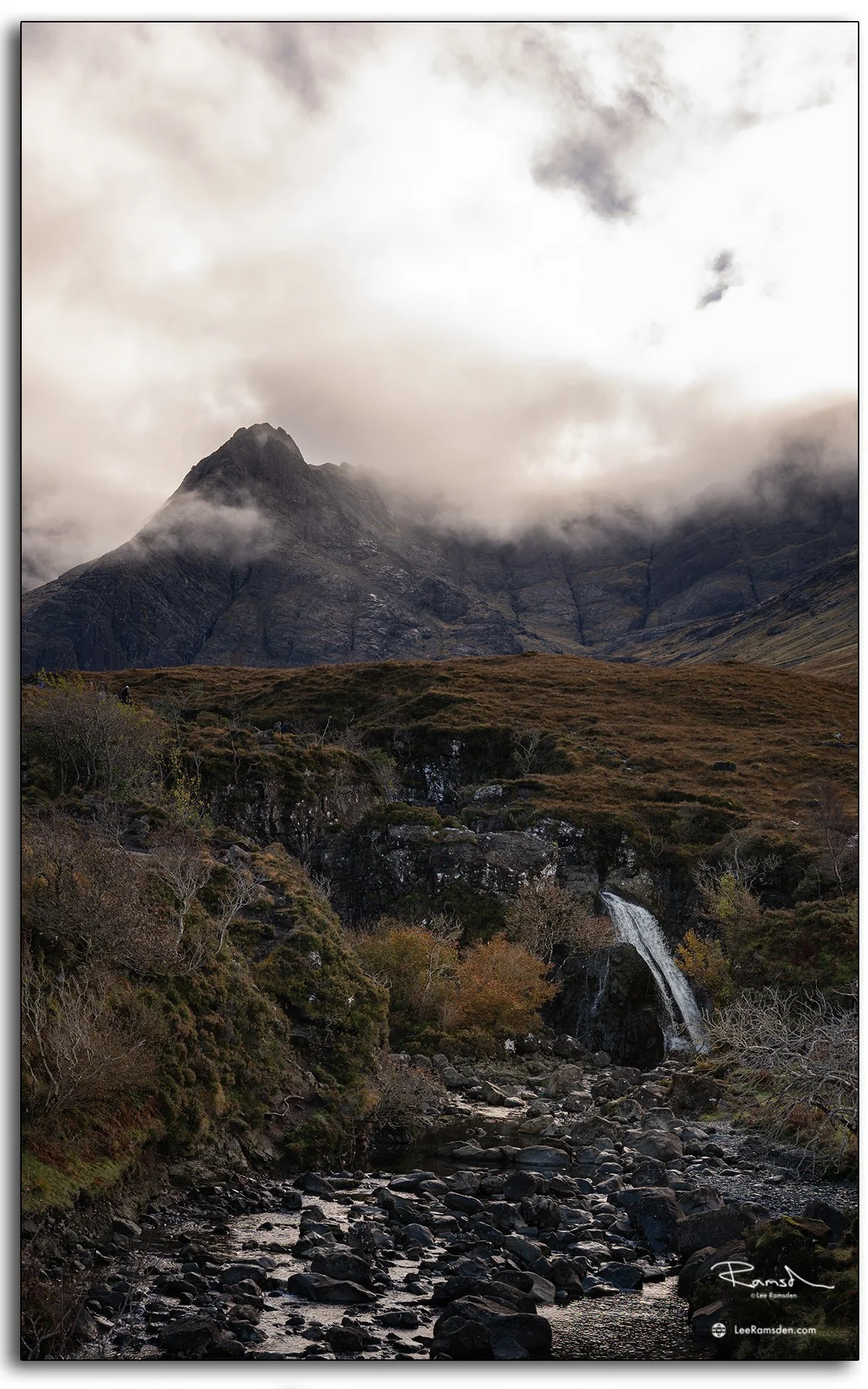 View towards the Cuillin mountains from the Fairy Pools on the Isle of Skye, with mist rolling over rugged peaks and a rocky stream in the foreground.