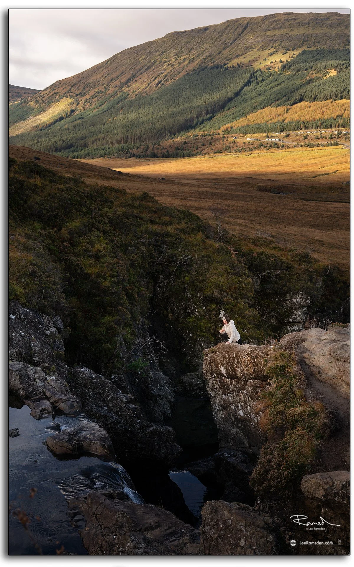 Woman sitting on a rocky ledge overlooking a waterfall at the Fairy Pools on the Isle of Skye, Scotland, surrounded by golden light and rugged hills