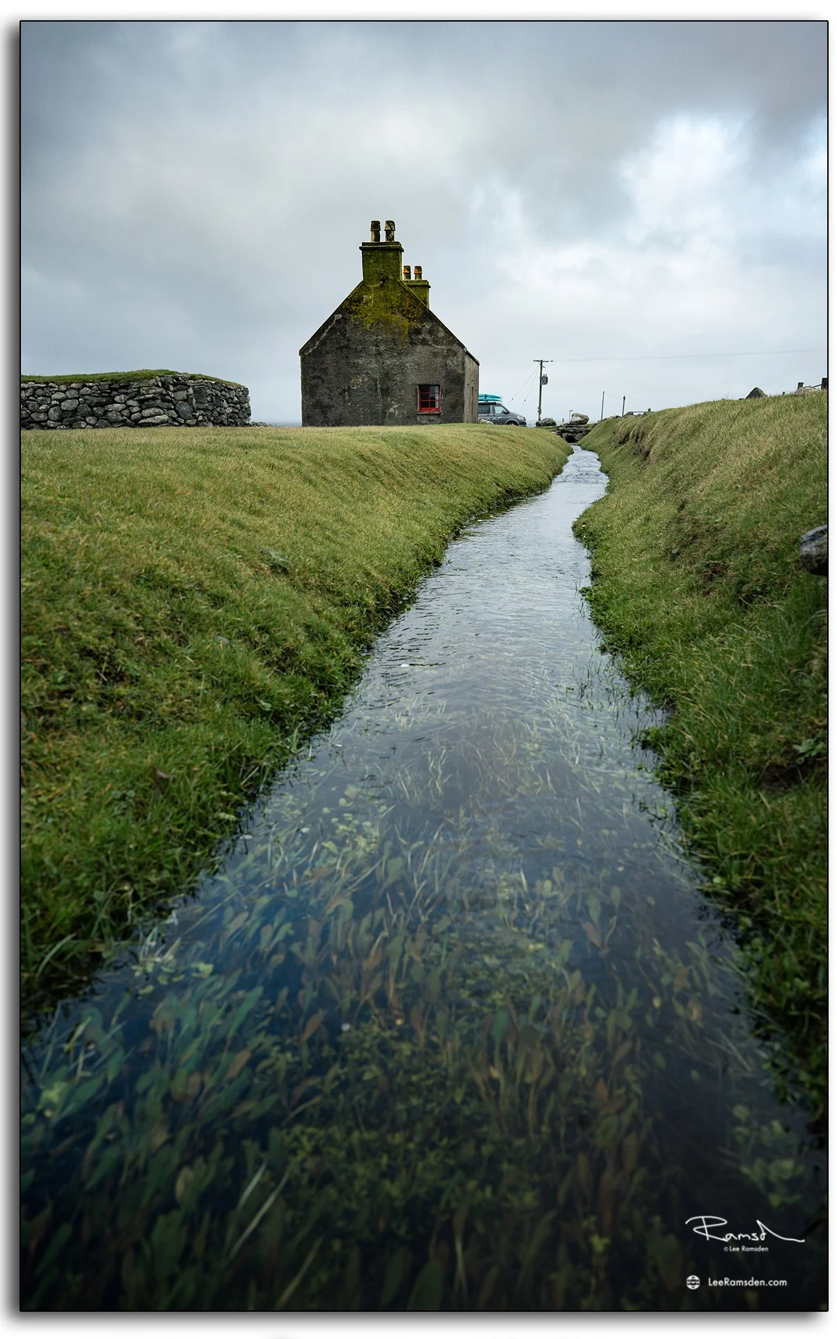 Historic blackhouse with moss-covered chimneys at Gearrannan on the Isle of Lewis, Scotland, viewed along a narrow grassy stream leading toward the sea.