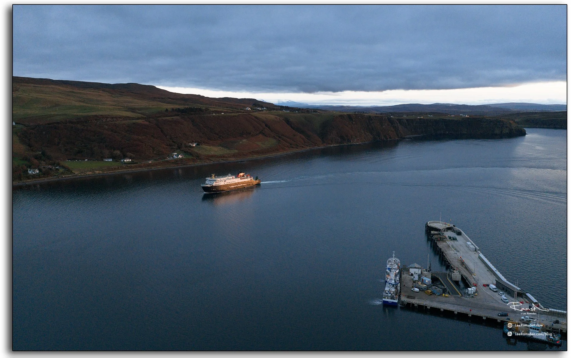 Aerial view overlooking Uig Harbour and village on the Isle of Skye, Scotland, with a camper van parked on the hillside and calm evening light over the bay