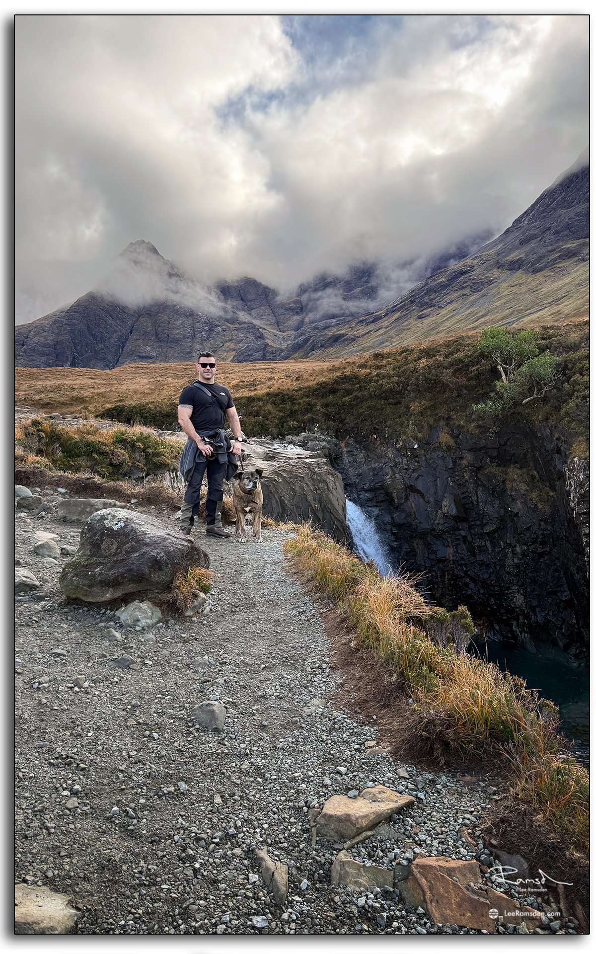 Man with his dog standing on a mountain trail beside a waterfall at the Fairy Pools, Isle of Skye, with dramatic clouds over the Cuillin mountains