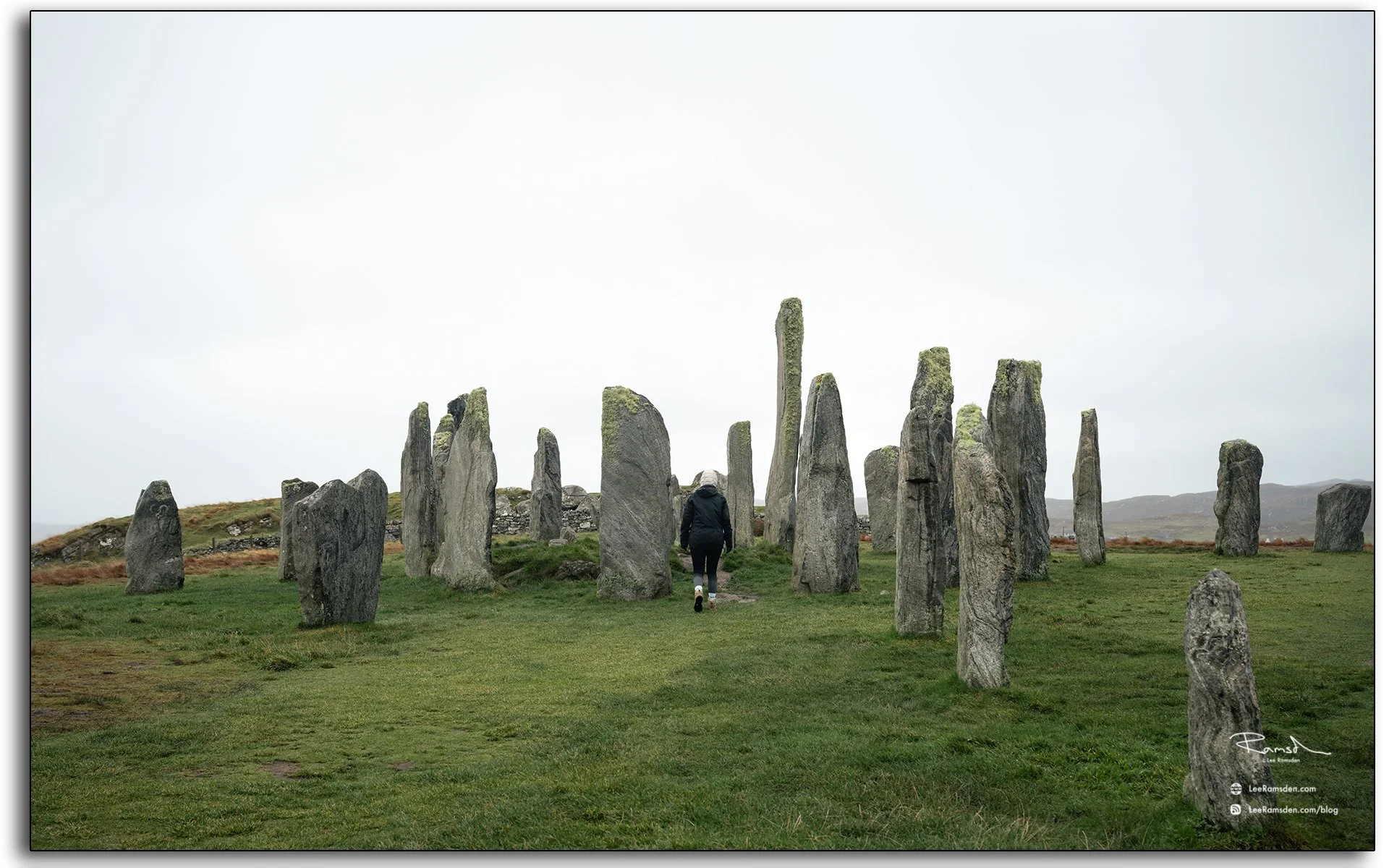 Person walking among the ancient Callanish Standing Stones on the Isle of Lewis, Scotland, surrounded by misty skies and rugged moorland