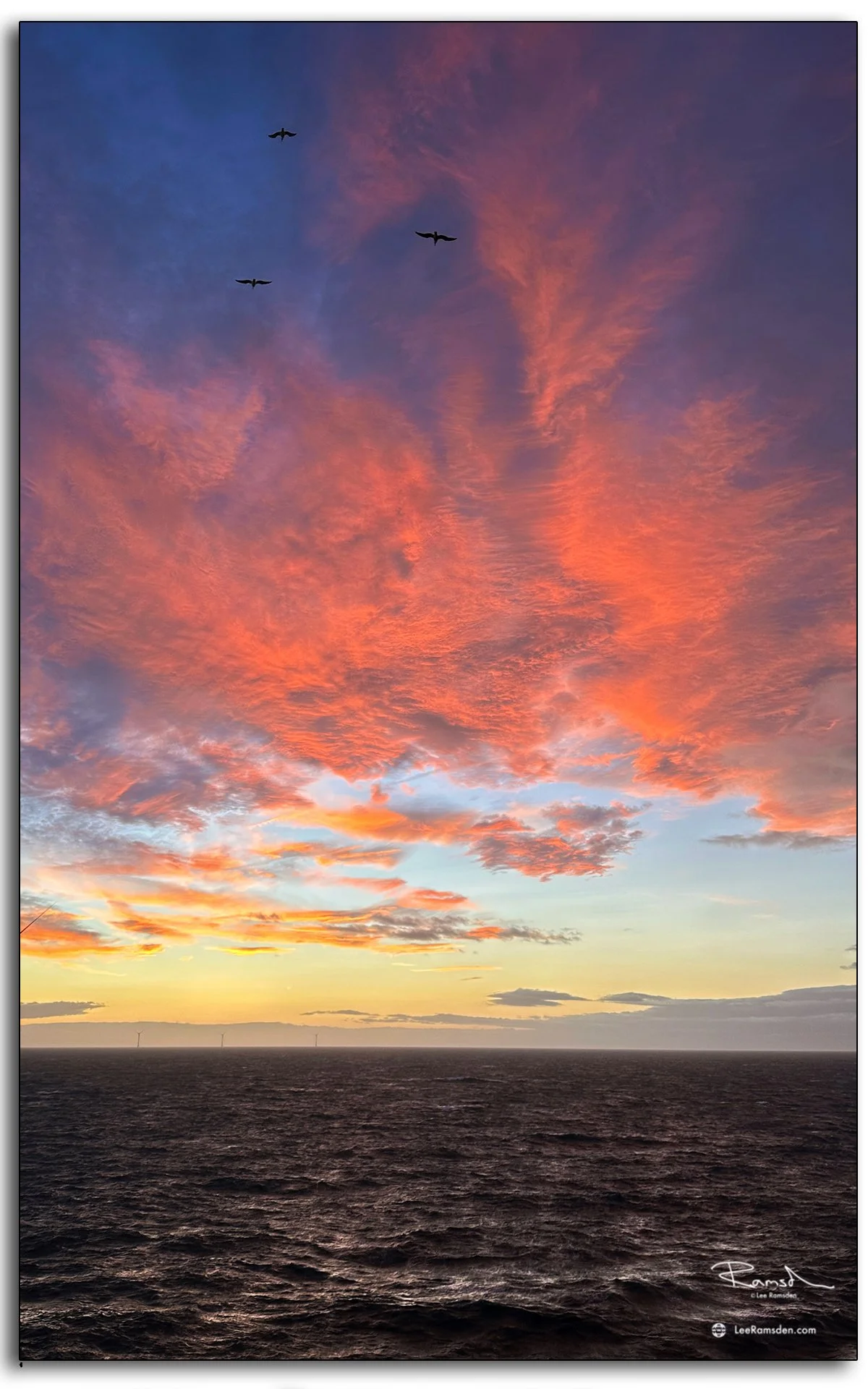 Row of offshore wind turbines lit by fiery orange clouds and evening light.