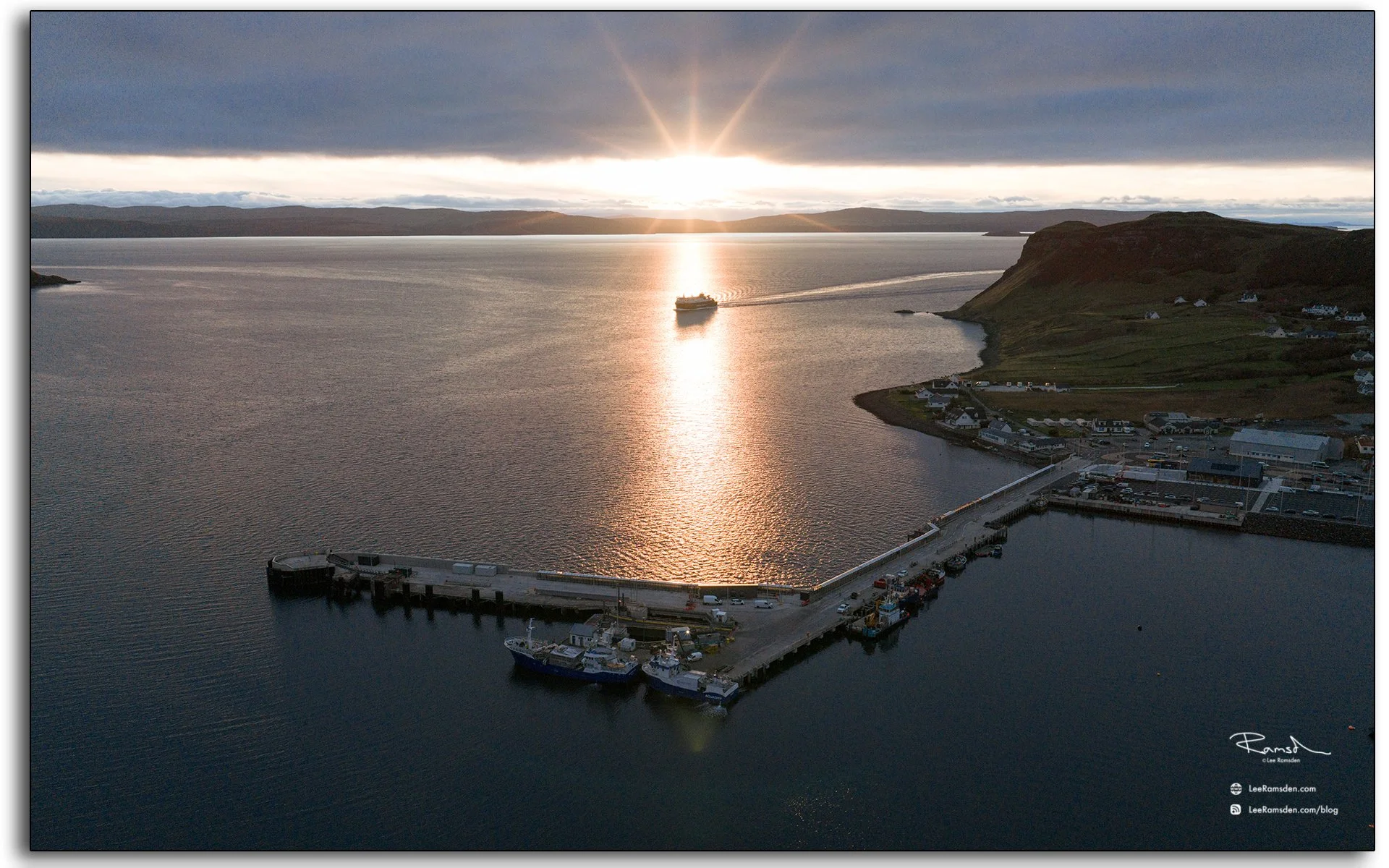 Aerial view of Uig Harbour on the Isle of Skye at sunset, with a ferry sailing across calm waters under a glowing orange sky