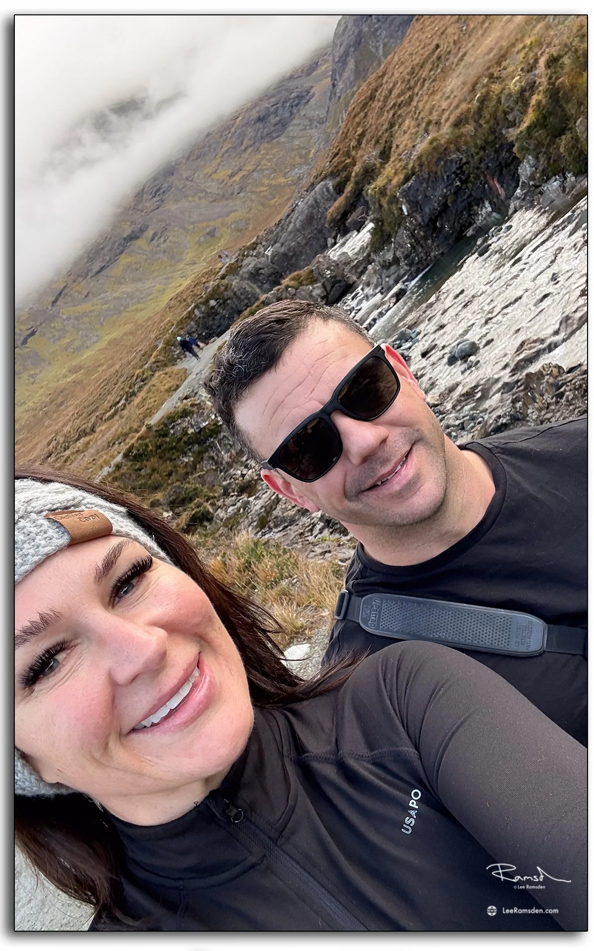 Couple taking a selfie during a hike at the Fairy Pools on the Isle of Skye, Scotland, with rugged cliffs and misty mountains in the background.
