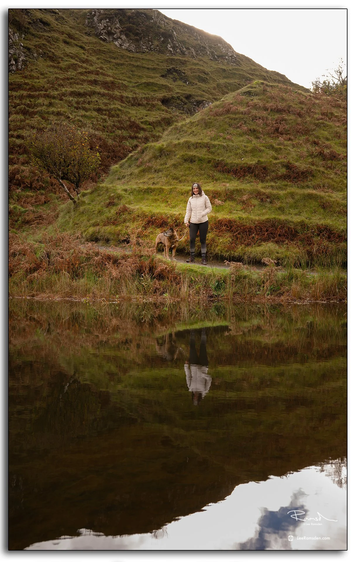 Woman walking beside a calm pond at the Fairy Glen on the Isle of Skye, with her reflection mirrored in the still water and grassy hills behind
