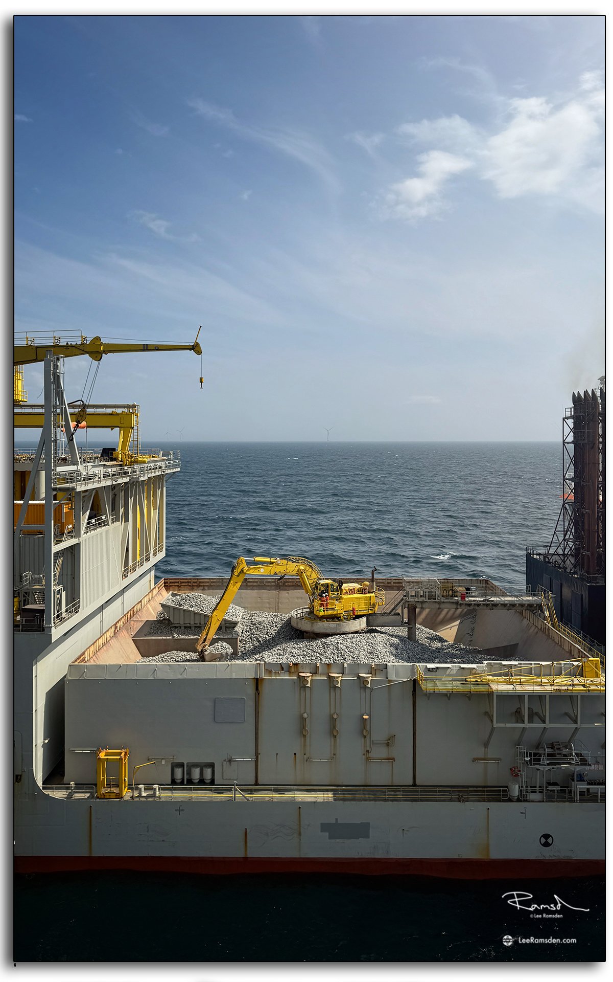 Mechanical digger working on rock load aboard Simon Stevin vessel, offshore wind farm backdrop