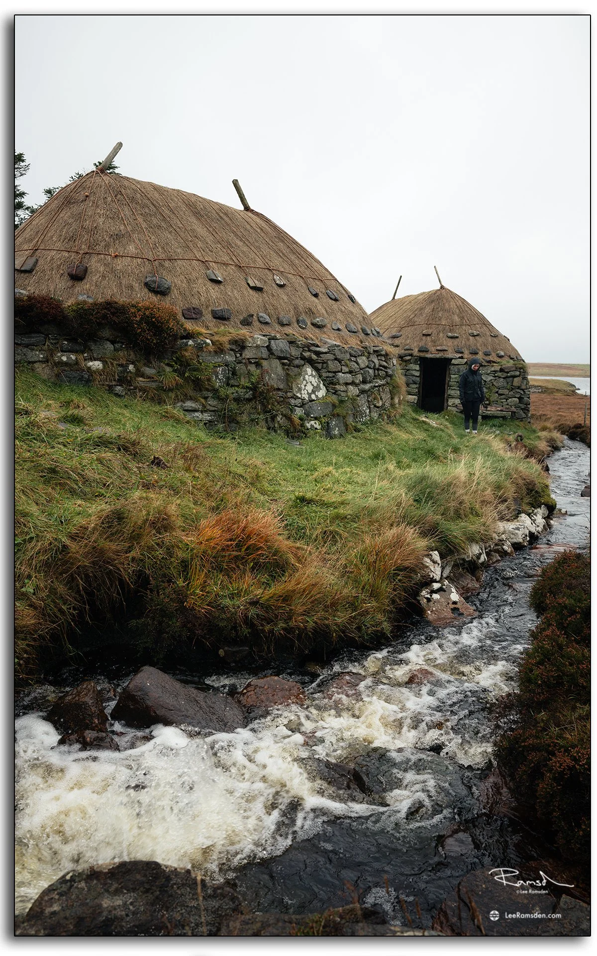 Traditional thatched blackhouses at Arnol on the Isle of Lewis, Scotland, with stone walls beside a flowing stream under grey Hebridean skies
