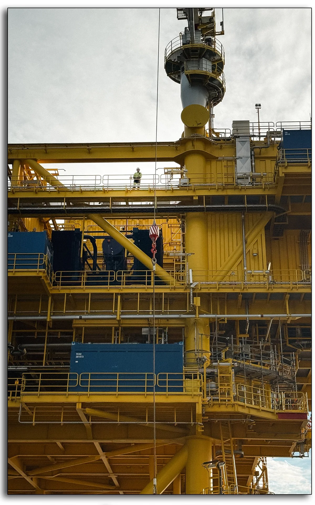 Worker on offshore substation platform in high-visibility PPE, safety harness, and helmet, standing on yellow structure, renewable energy industry.
