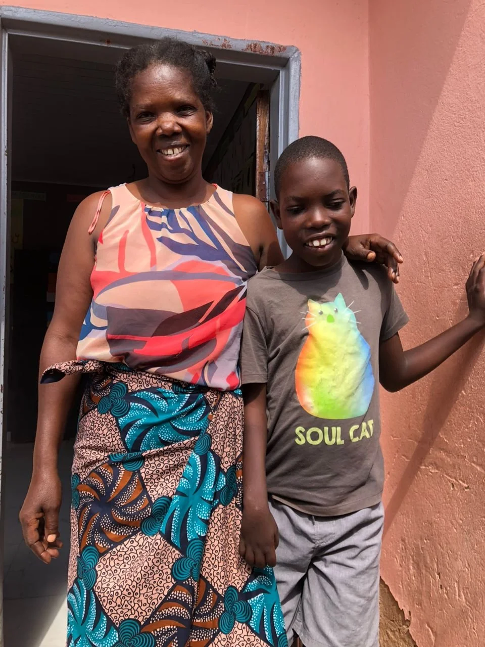 A Zambian mother in a colorful dress stands with her arm around her son wearing a grey t-shirt with a colorful cat displayed.